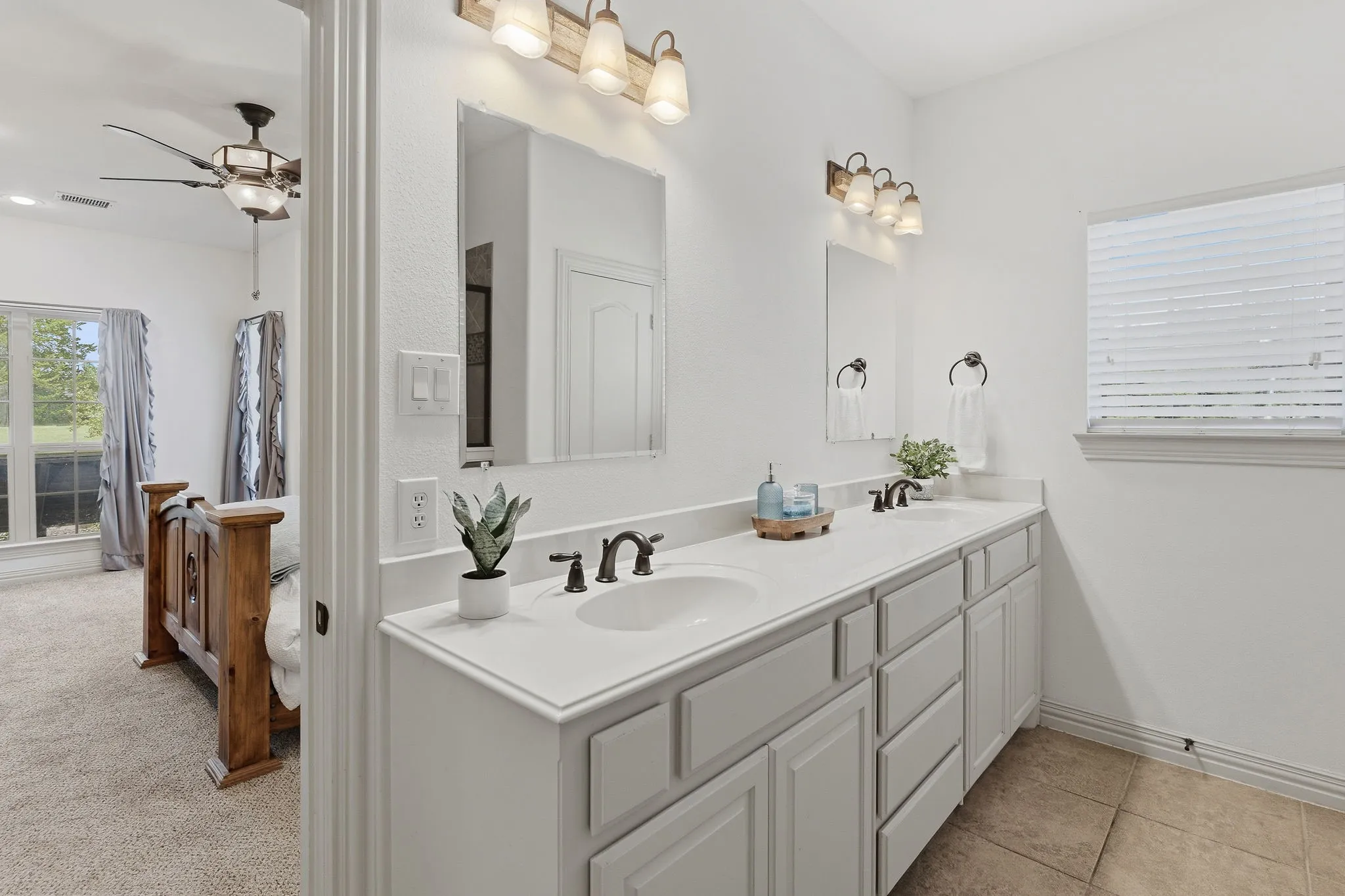 The primary bath combines an extended dual-sink vanity, generous counter space, abundant white cabinetry, and individual mirrors with warm tile flooring, bronze-tone fixtures, and natural light for a bright, inviting space to begin and end each day.