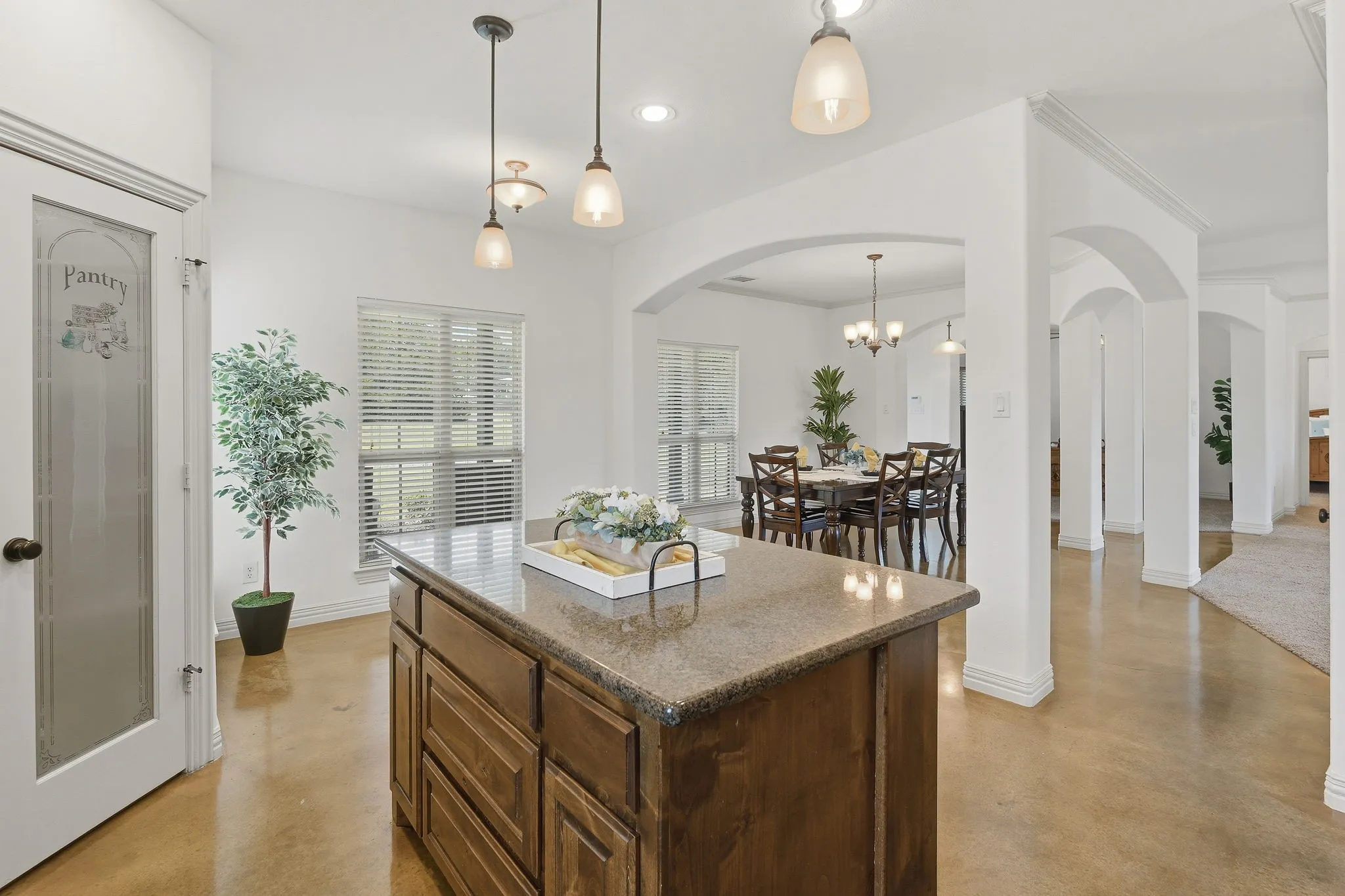 A spacious island beneath pendant lighting anchors the kitchen, with a decorative glass pantry door, stained concrete flooring, and graceful archways connecting the nearby dining and living spaces for easy entertaining.