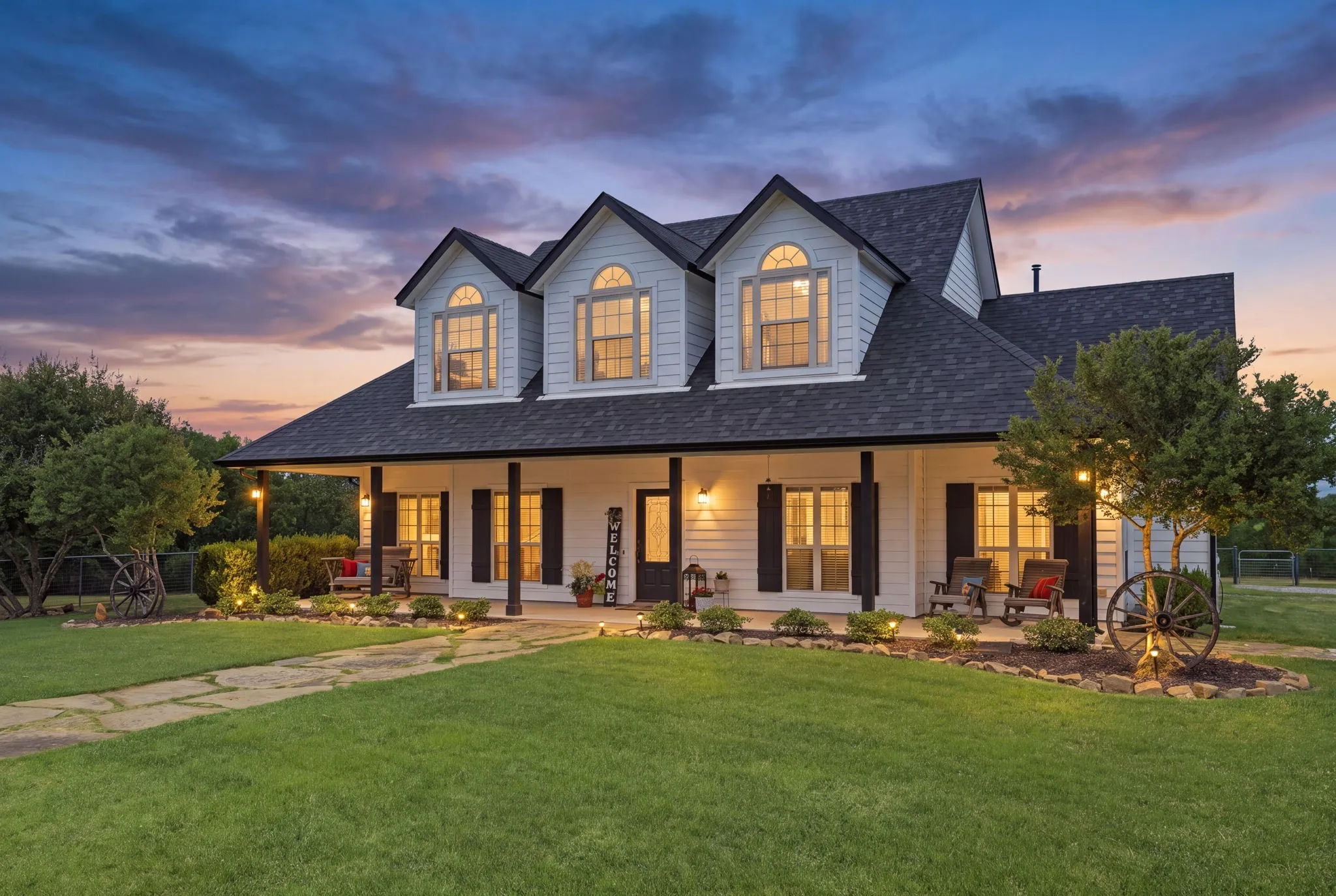 Warmly glowing windows, arched dormers, and softly illuminated landscaping showcase the farmhouse’s inviting character beneath a colorful evening sky.