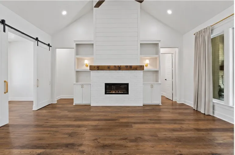 Vaulted ceiling, wood-finish flooring, white brick fireplace with wood mantel, built-in shelving, and barn door entry