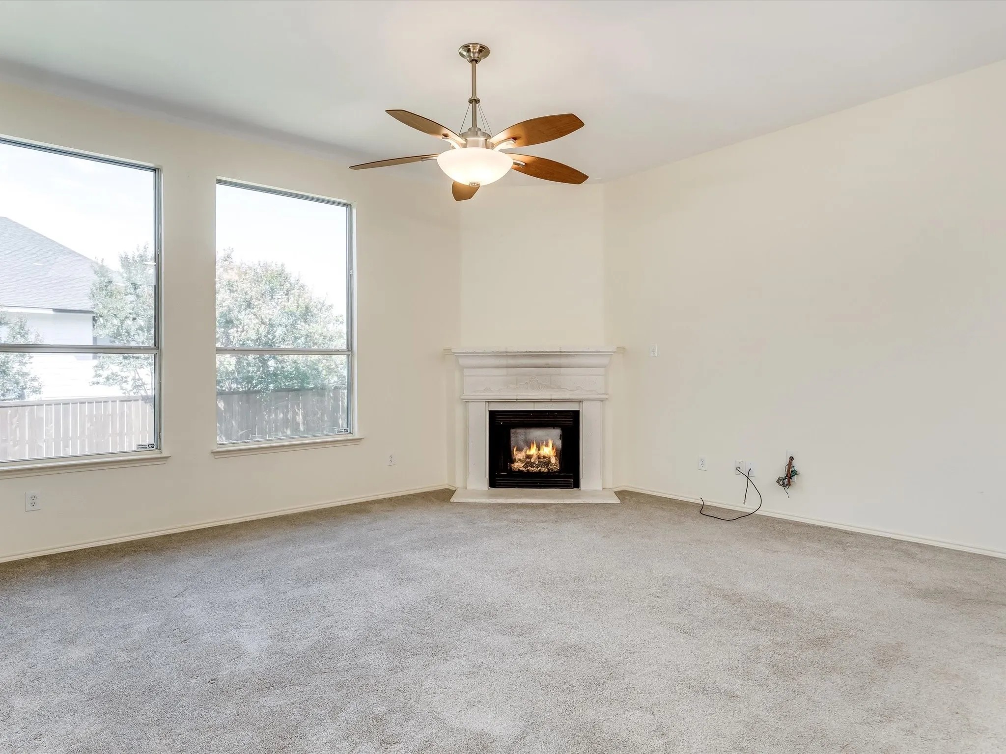 Spacious interior room featuring a corner fireplace with a decorative mantel, two large windows, a ceiling fan with integrated lighting, and light-colored carpeting