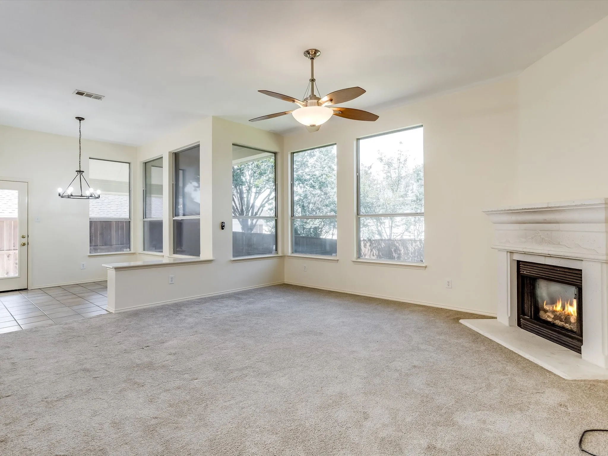 Spacious room featuring a fireplace with a stone mantel, multiple large windows, a ceiling fan with integrated lighting, and carpet flooring
