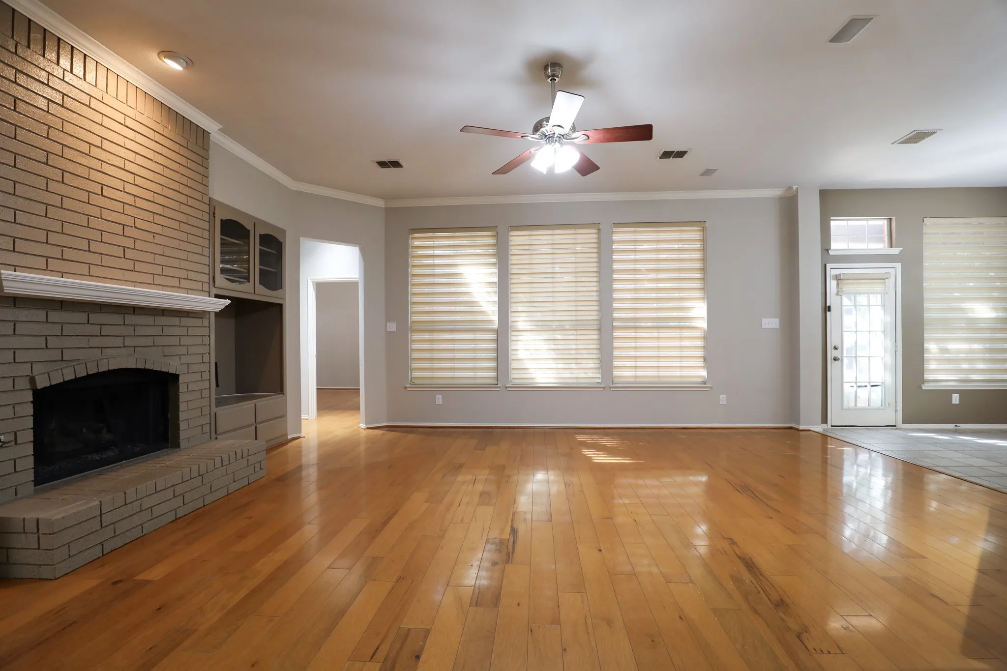 Expansive living area featuring wood-finish flooring, a brick fireplace with a mantel, built-in cabinetry, and multiple windows with blinds