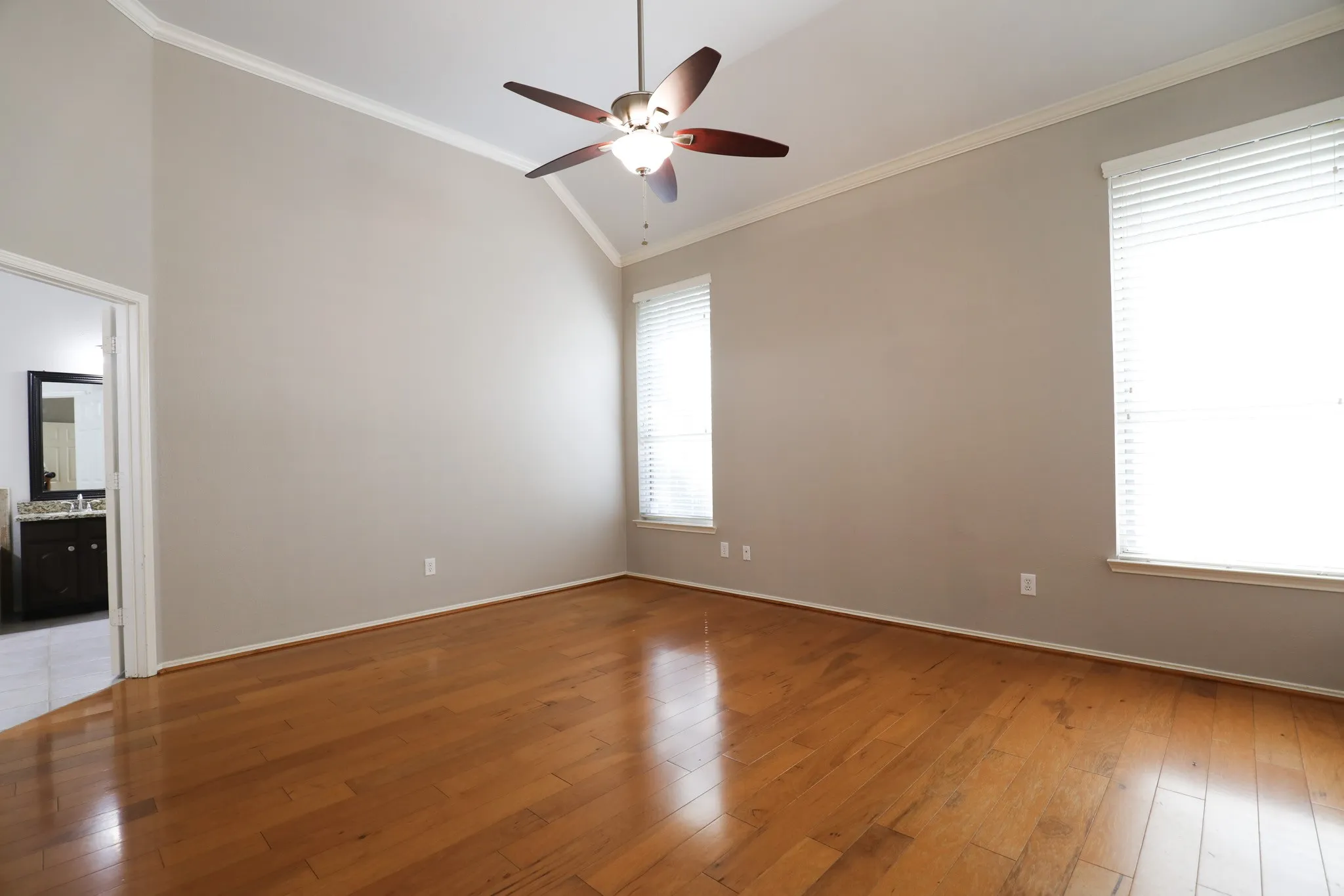 Vaulted ceiling with crown molding, wood-finish flooring, two large windows with blinds, and a ceiling fan with integrated lighting