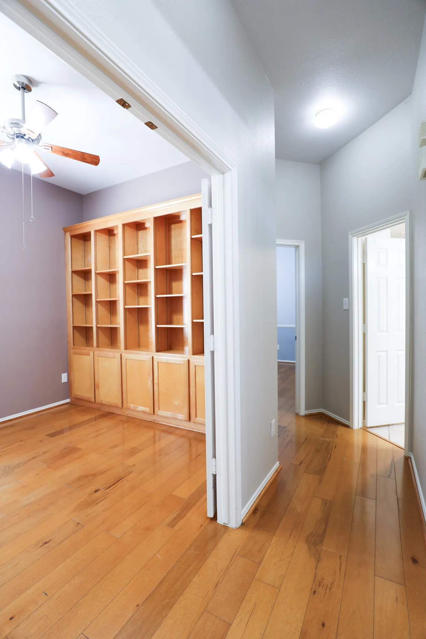 Wood-finish flooring throughout, built-in shelving with lower cabinetry, ceiling fan with light fixture, and recessed lighting in the hallway