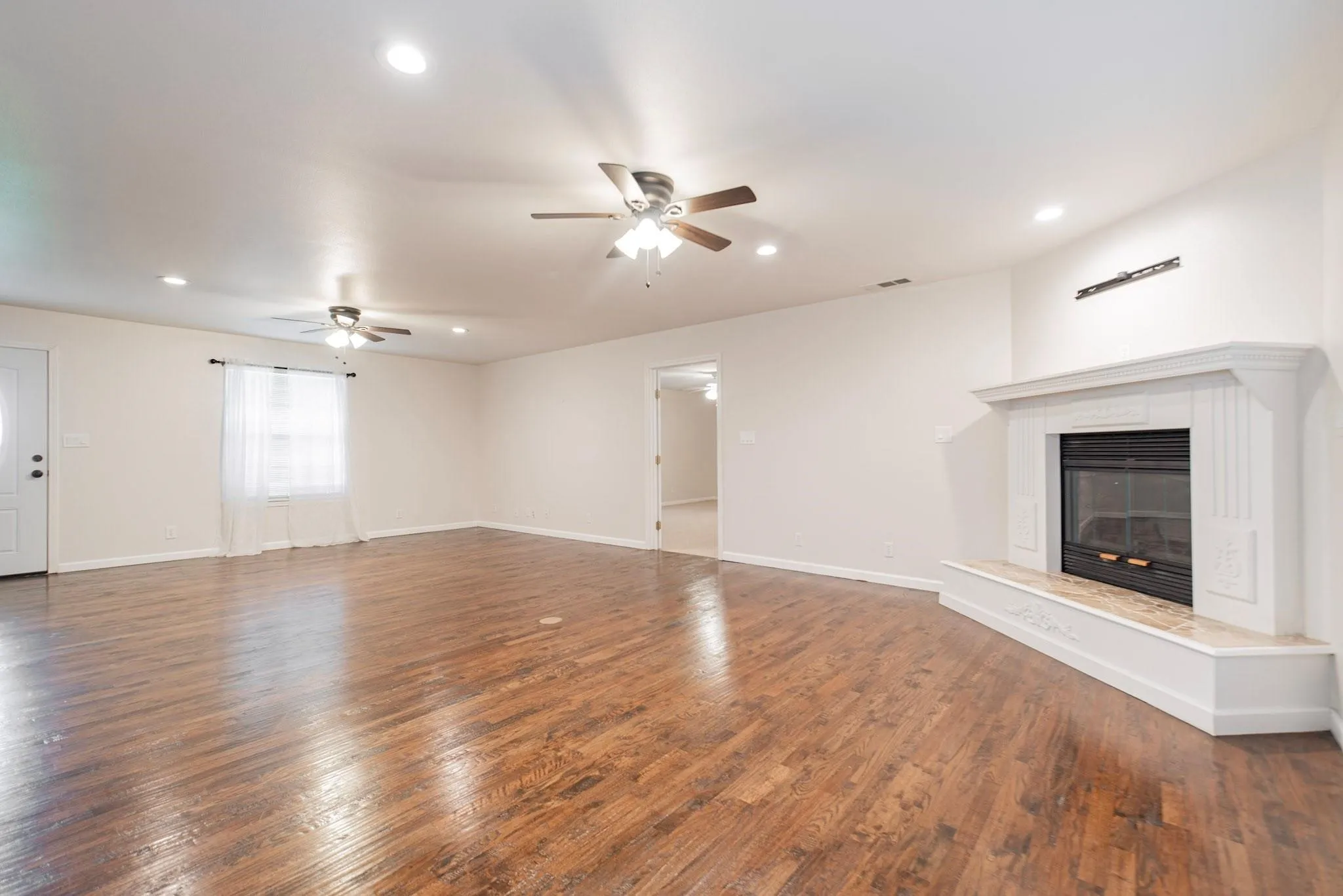 Spacious living area featuring wood-finish flooring, a white mantel fireplace with a hearth, multiple ceiling fans, recessed lighting, and a white entry door