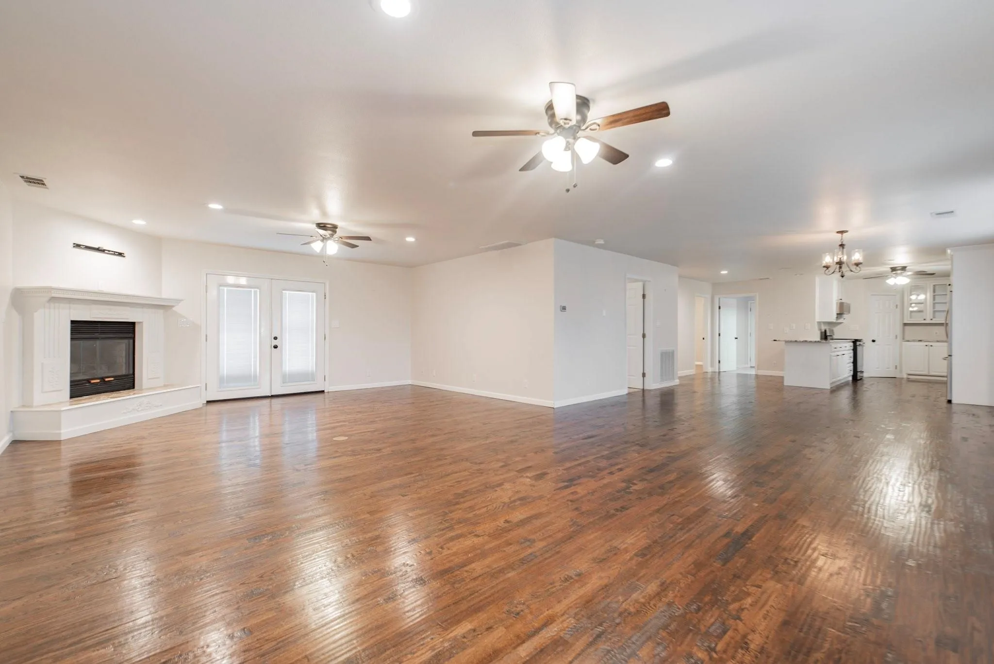 Expansive interior space featuring wood-finish flooring, a white mantel fireplace, double glass-paneled doors, recessed lighting, and ceiling fans
