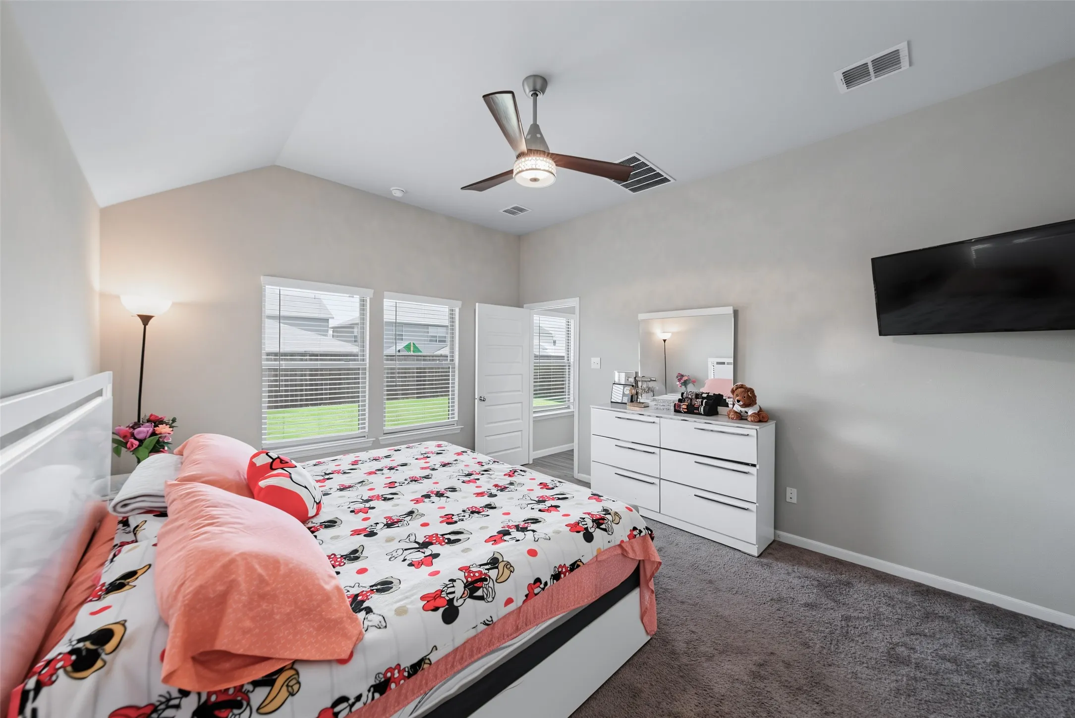 Vaulted ceiling, ceiling fan with wood-finish blades, three large windows with blinds, gray wall paint, and gray carpet flooring