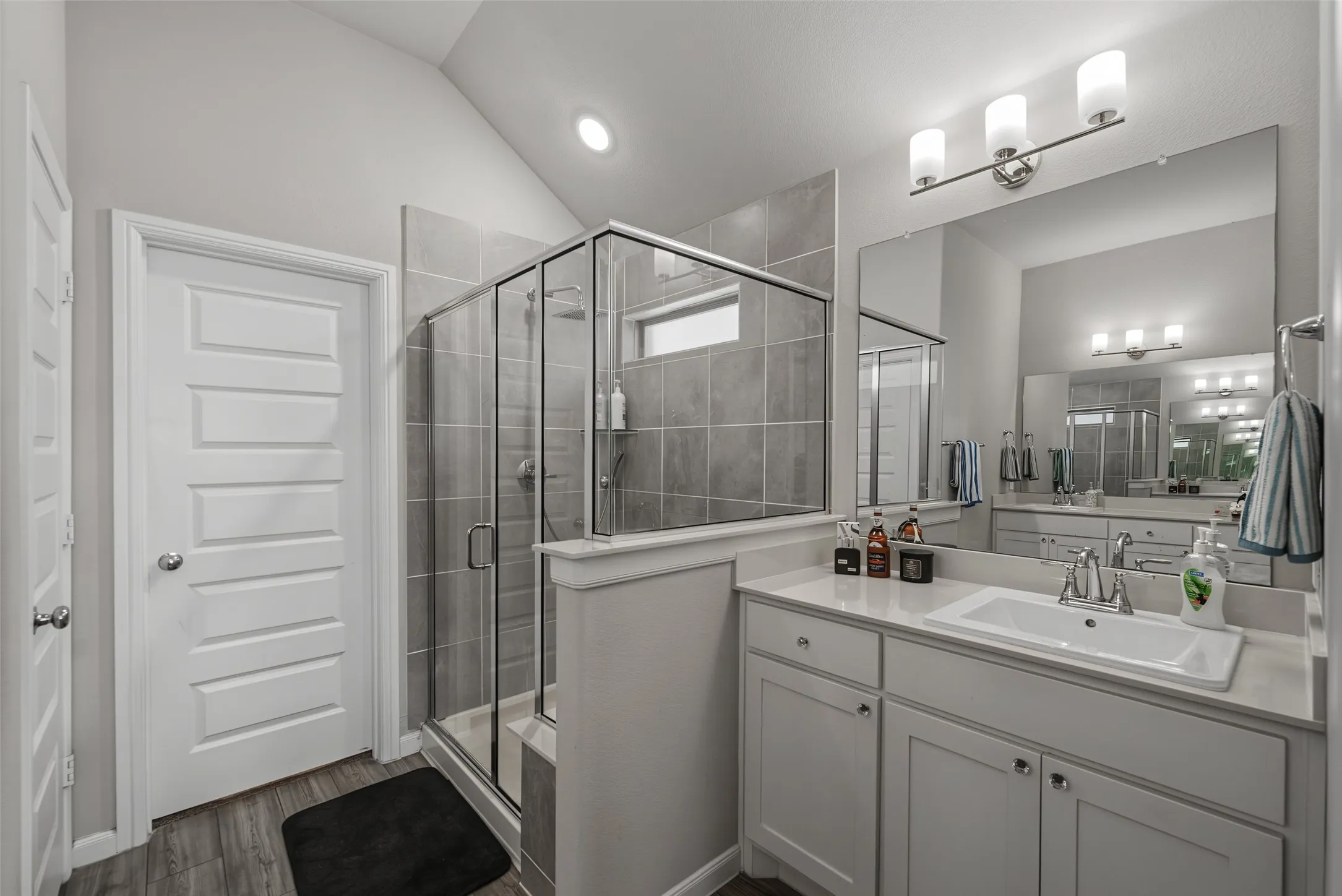 Glass-enclosed shower with gray tile surround, wood-finish flooring, white vanity with integrated sink, and large wall-mounted mirror