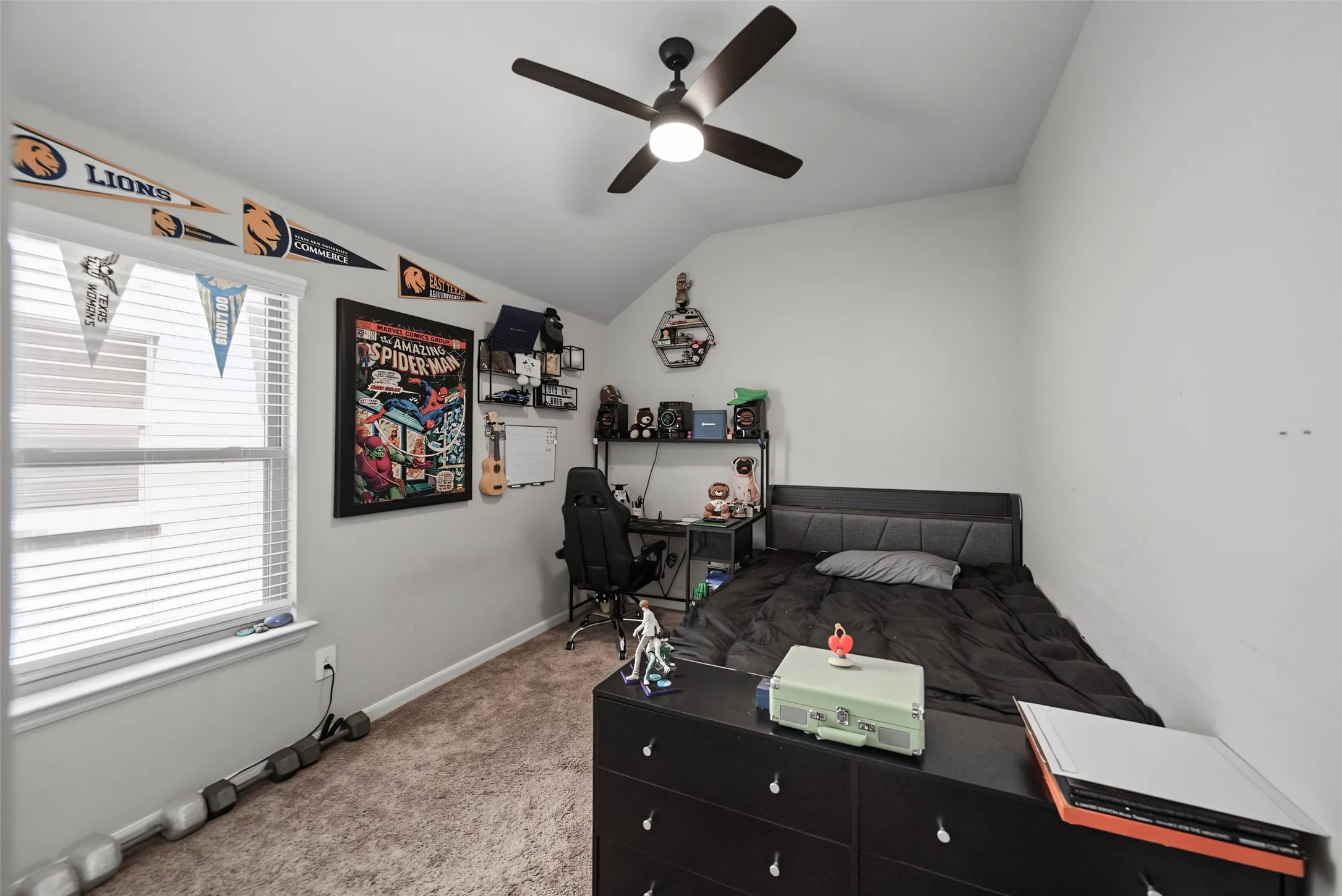 Carpeted flooring, vaulted ceiling, window with blinds, ceiling fan with integrated lighting, and a built-in wall shelf