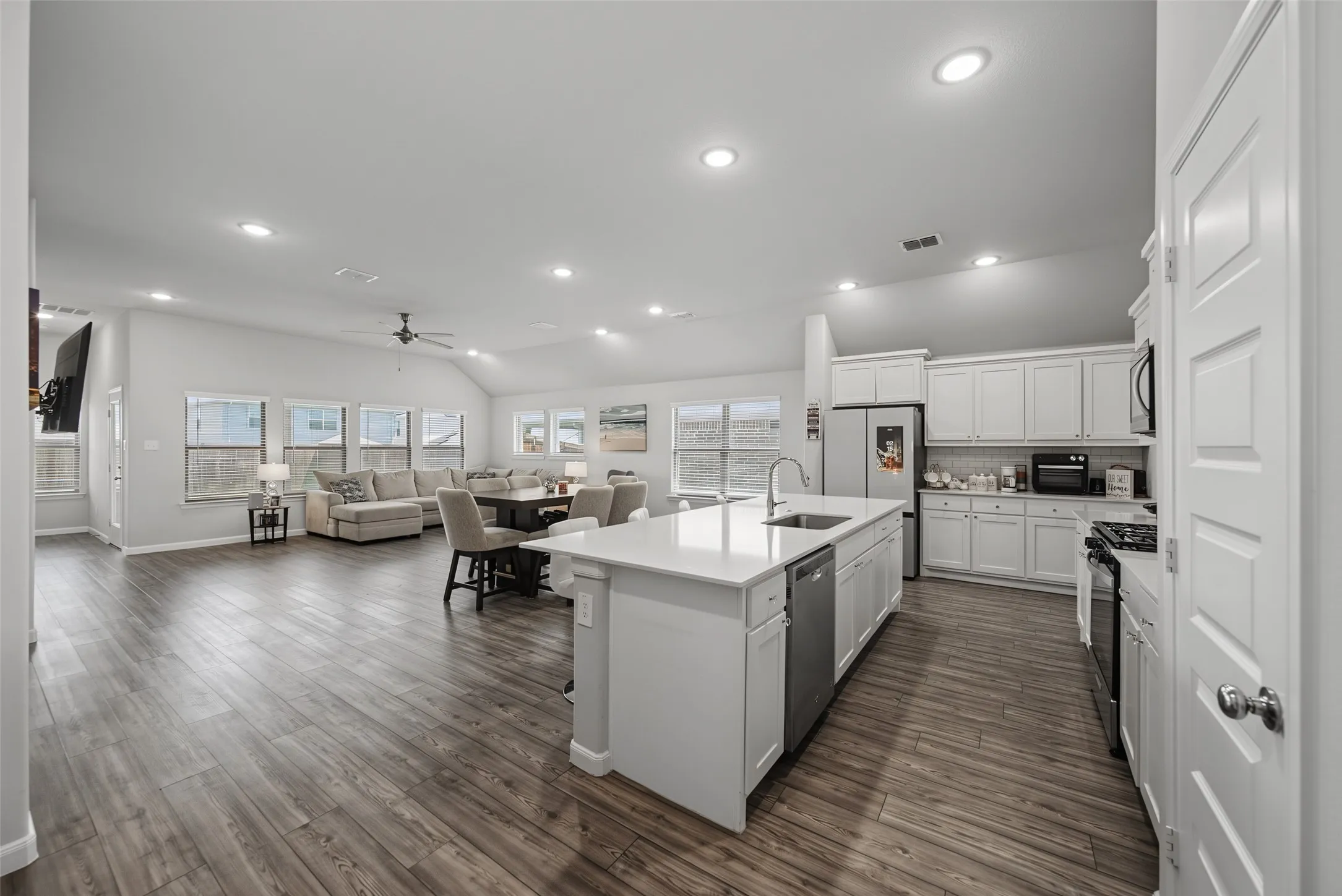 Open-concept living area featuring wood-finish flooring, recessed lighting, a vaulted ceiling, a spacious kitchen island with an integrated sink, and white cabinetry