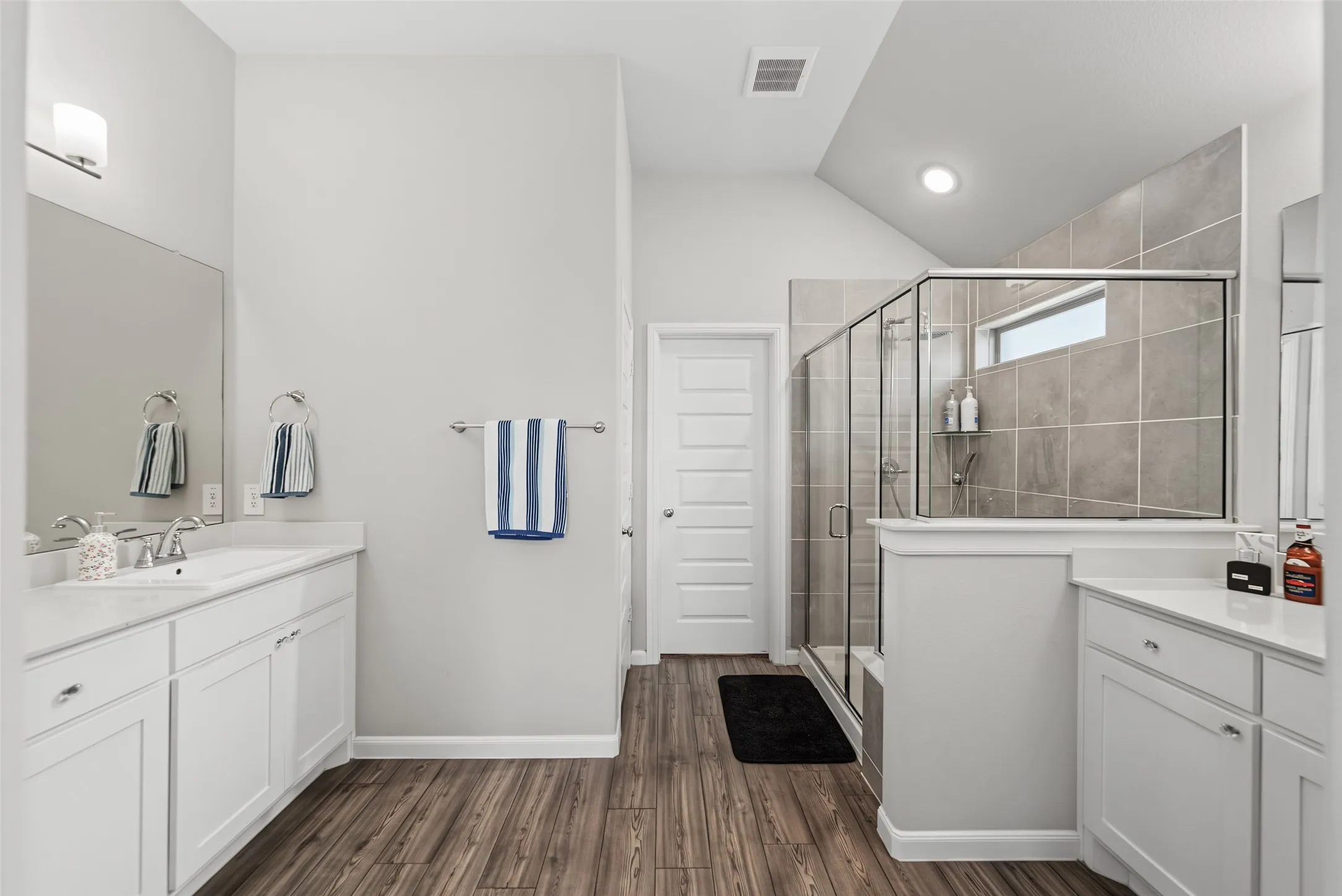 Dual vanity with white cabinetry, wood-finish flooring, and a glass-enclosed shower with tile surround
