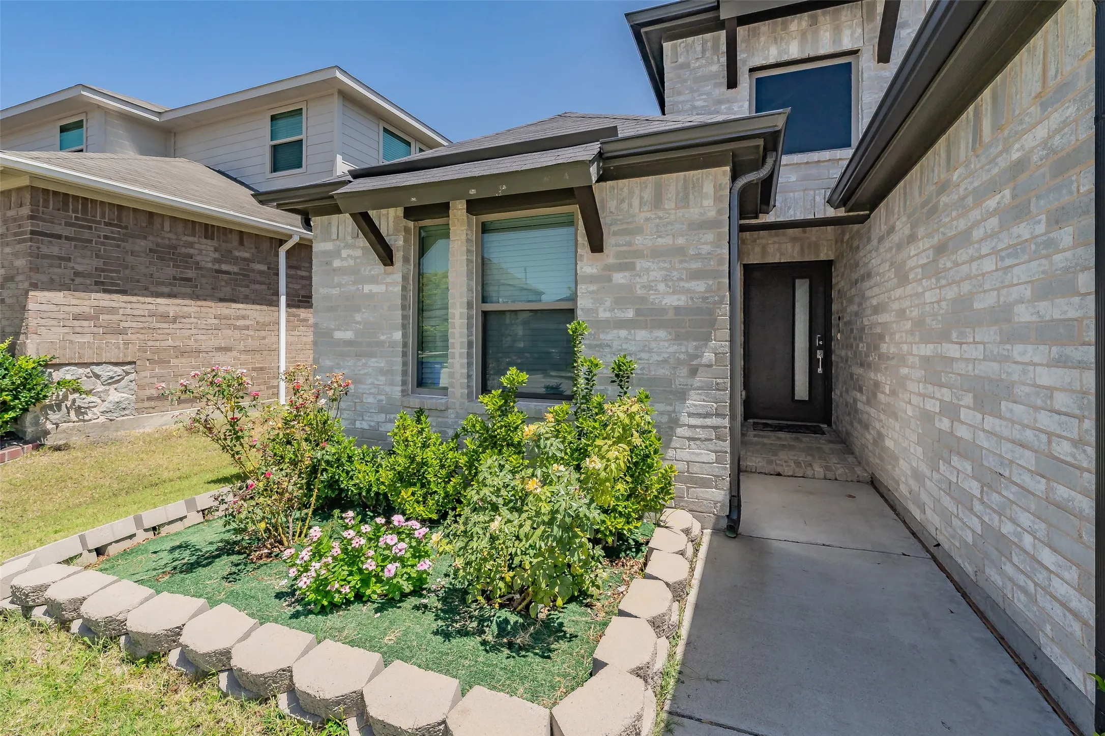 Modern brick exterior with contemporary architectural detailing, featuring large multi-pane windows, a covered entry walkway with decorative pavers, and mature landscaping including ornamental shrubs and established lawn areas