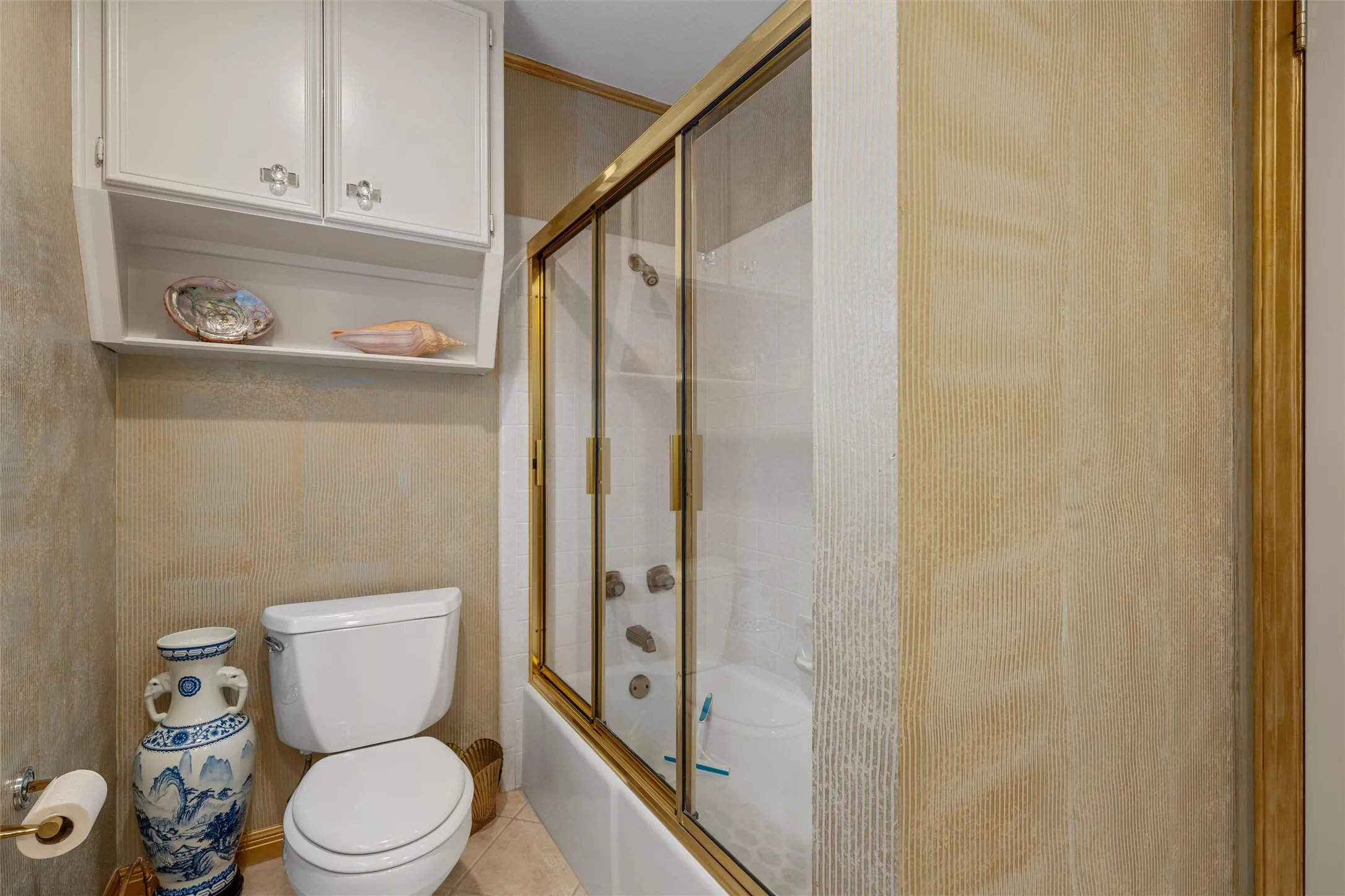 Bathroom featuring an enclosed shower-tub combination with gold-tone framing, white cabinetry with clear hardware, a two-piece toilet, and textured wall finishes