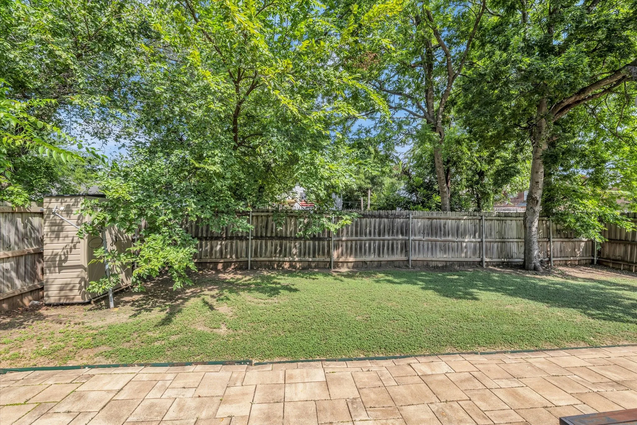 Expansive backyard featuring a stone paver patio, a manicured lawn, a wood privacy fence, and mature trees providing natural shade