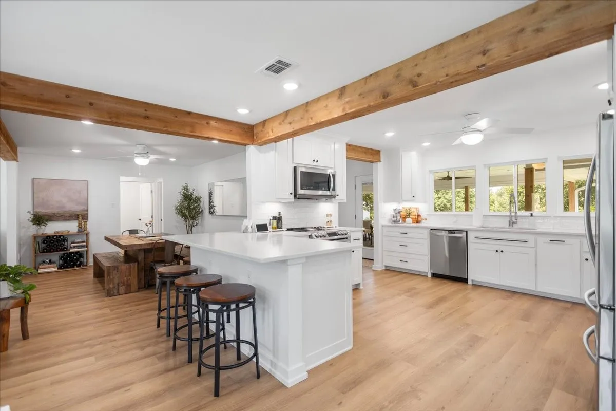 Open-concept kitchen featuring wood-finish flooring, exposed wood beams, crisp white custom cabinetry, integrated stainless steel appliances, and a central island with countertop seating