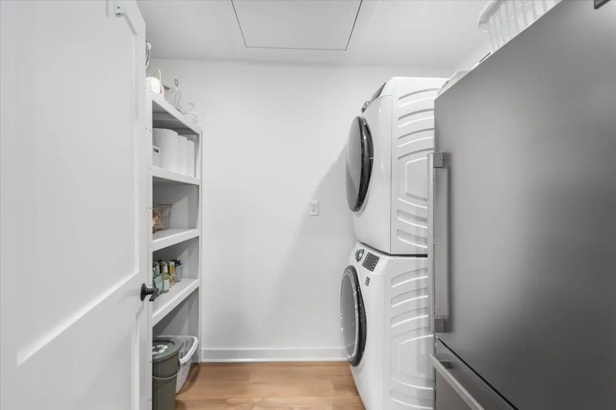 Dedicated laundry area featuring a stacked washer and dryer unit, wood-finish flooring, and shelving