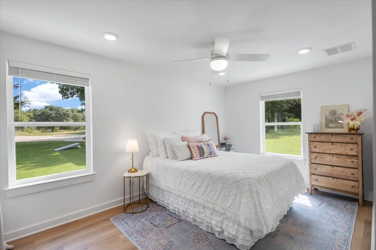 Bright Guest bedroom featuring expansive windows with pastoral outlooks, hardwood flooring, ceiling fan with integrated lighting, wood-finish dresser with multiple drawers, bedside table lamp, and area rug