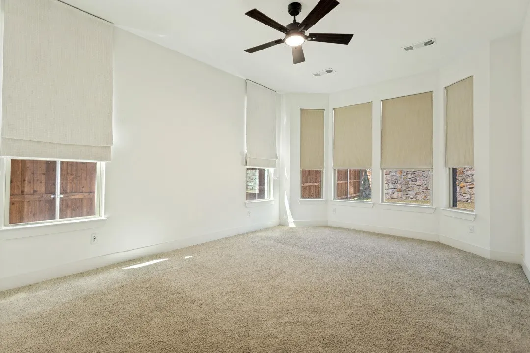 Bright, sun-filled room featuring expansive windows with cellular shades, neutral carpeting, and a modern ceiling fan with integrated lighting