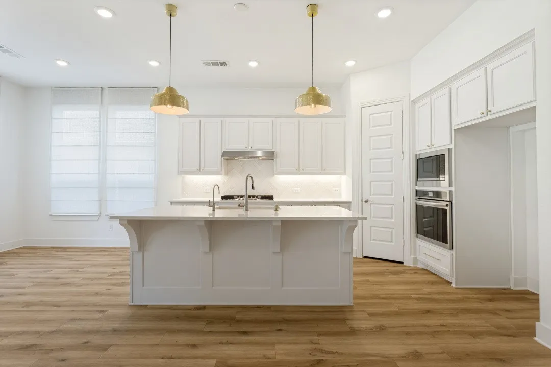 Central kitchen island with an undermount sink, white cabinetry, integrated stainless steel appliances, wood-finish flooring, and two pendant light fixtures