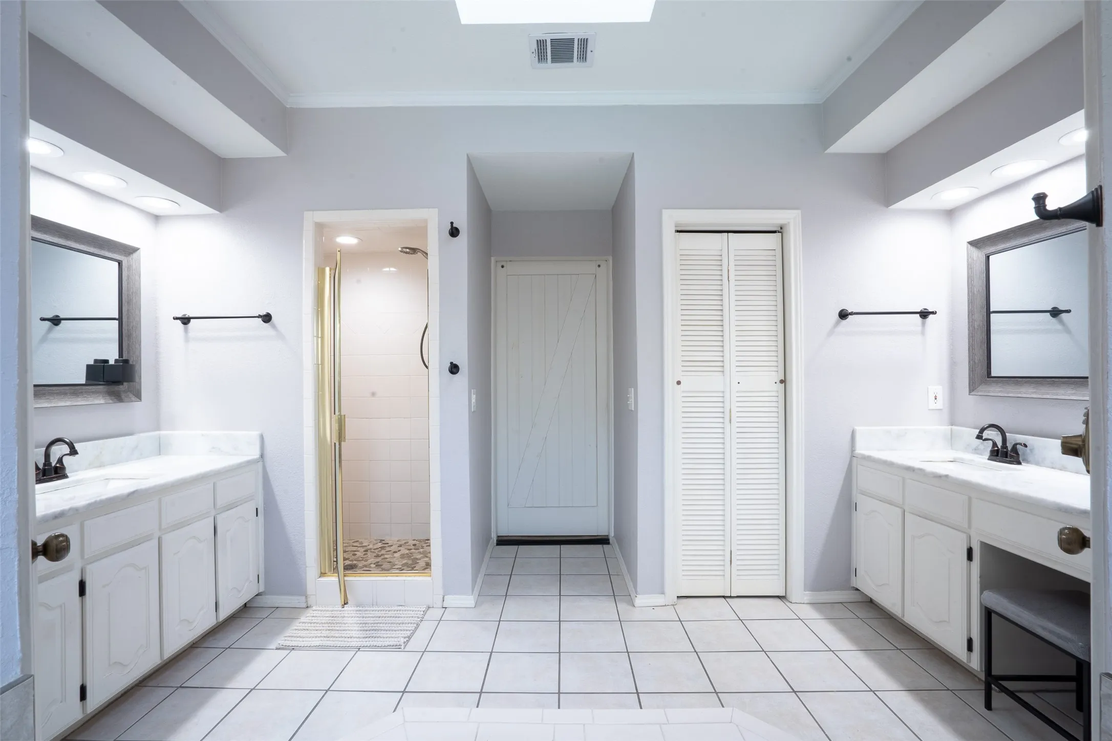 Bathroom featuring dual vanity areas with white cabinetry and light-toned countertops, a glass-enclosed shower with pebble-finish flooring, white tiled flooring, and a skylight