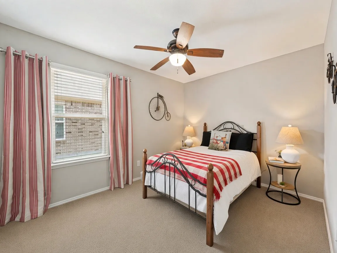 Comfortable secondary bedroom featuring a ceiling fan with integrated lighting, neutral carpet flooring, and large window with striped curtains and plantation shutters