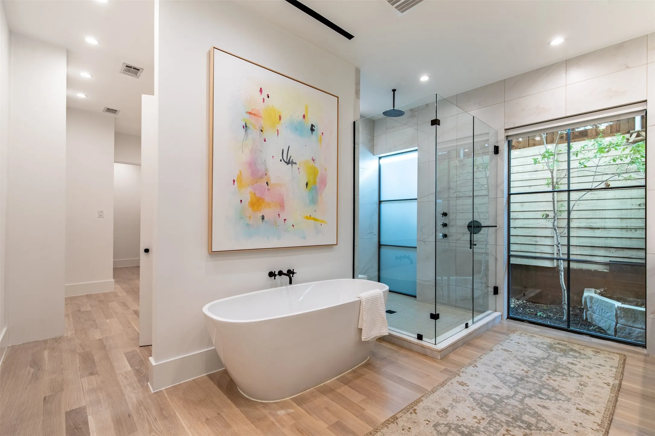 Freestanding soaking tub with matte black fixtures, a spacious glass-enclosed shower, and a large window providing natural light