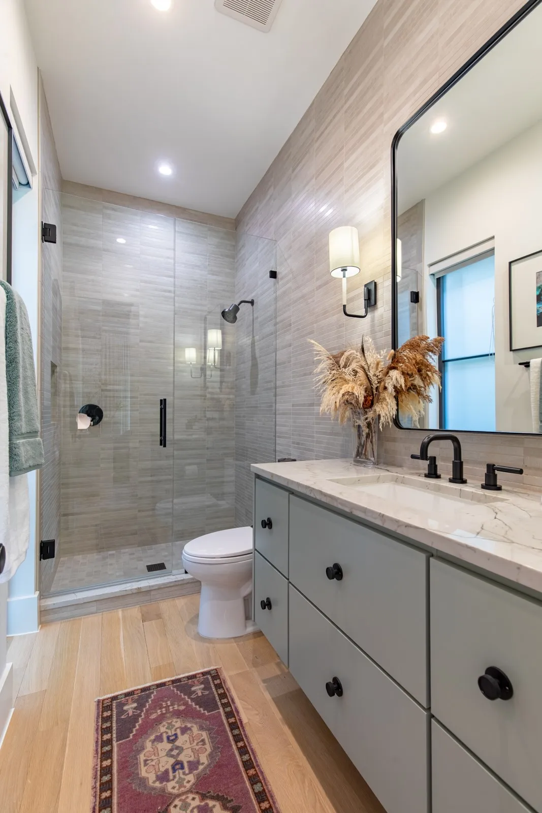 Modern bathroom featuring a glass-enclosed shower with tile surround, light wood-finish flooring, a double vanity with light gray cabinetry, a white countertop, and a large wall-mounted mirror