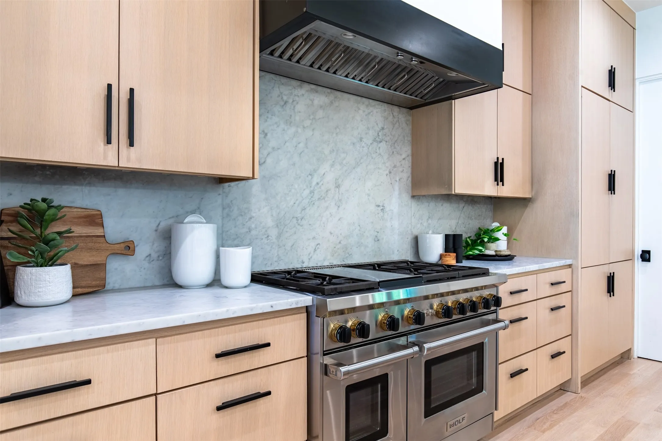 Gourmet kitchen featuring a professional-grade stainless steel range with a ventilation hood, light wood-finish cabinetry, black hardware, a stone backsplash, and white countertops