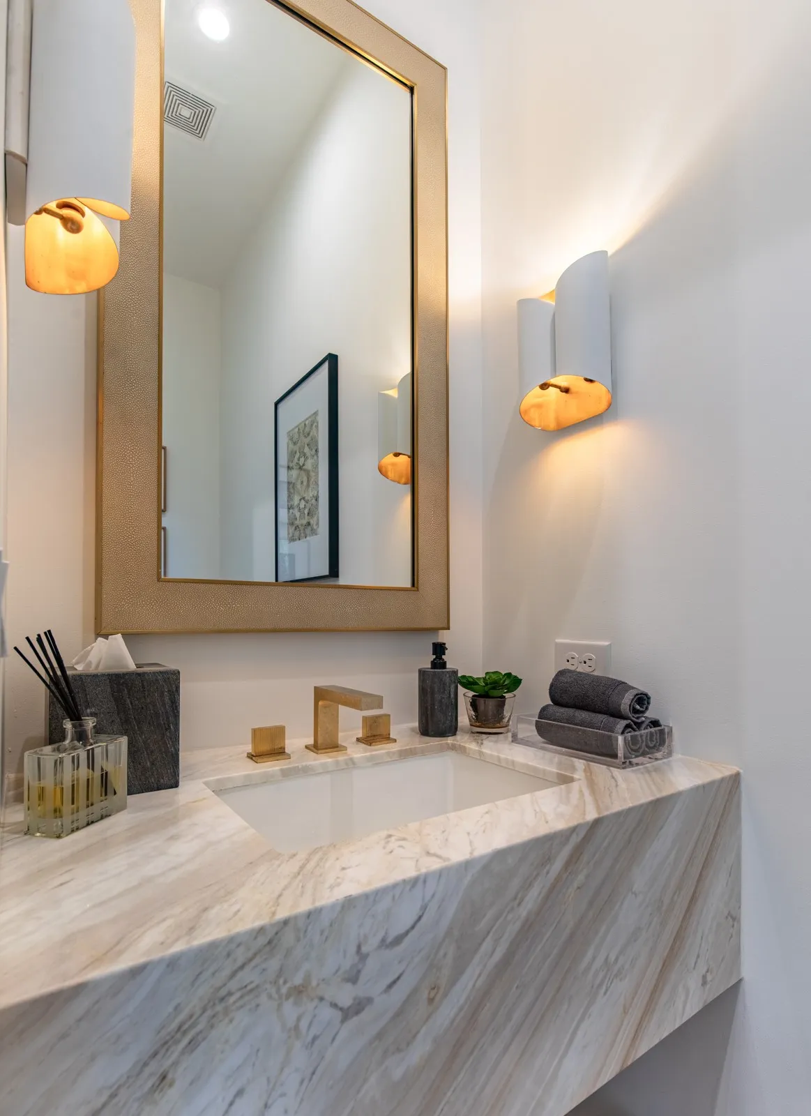 Floating vanity with a stone countertop and integrated sink, complemented by a large framed mirror, wall-mounted sconces, and a brushed gold faucet
