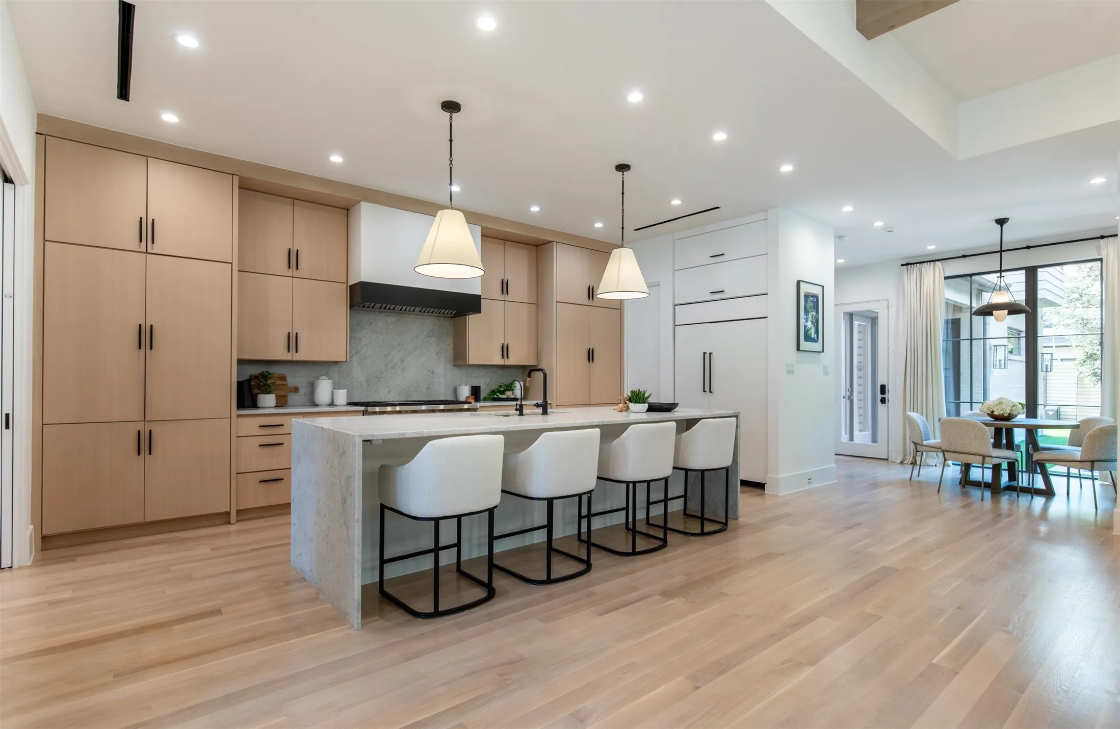 Open-concept kitchen featuring light wood-finish flooring, a waterfall island with a light-toned countertop, integrated appliances, recessed lighting, and a dining area with large windows