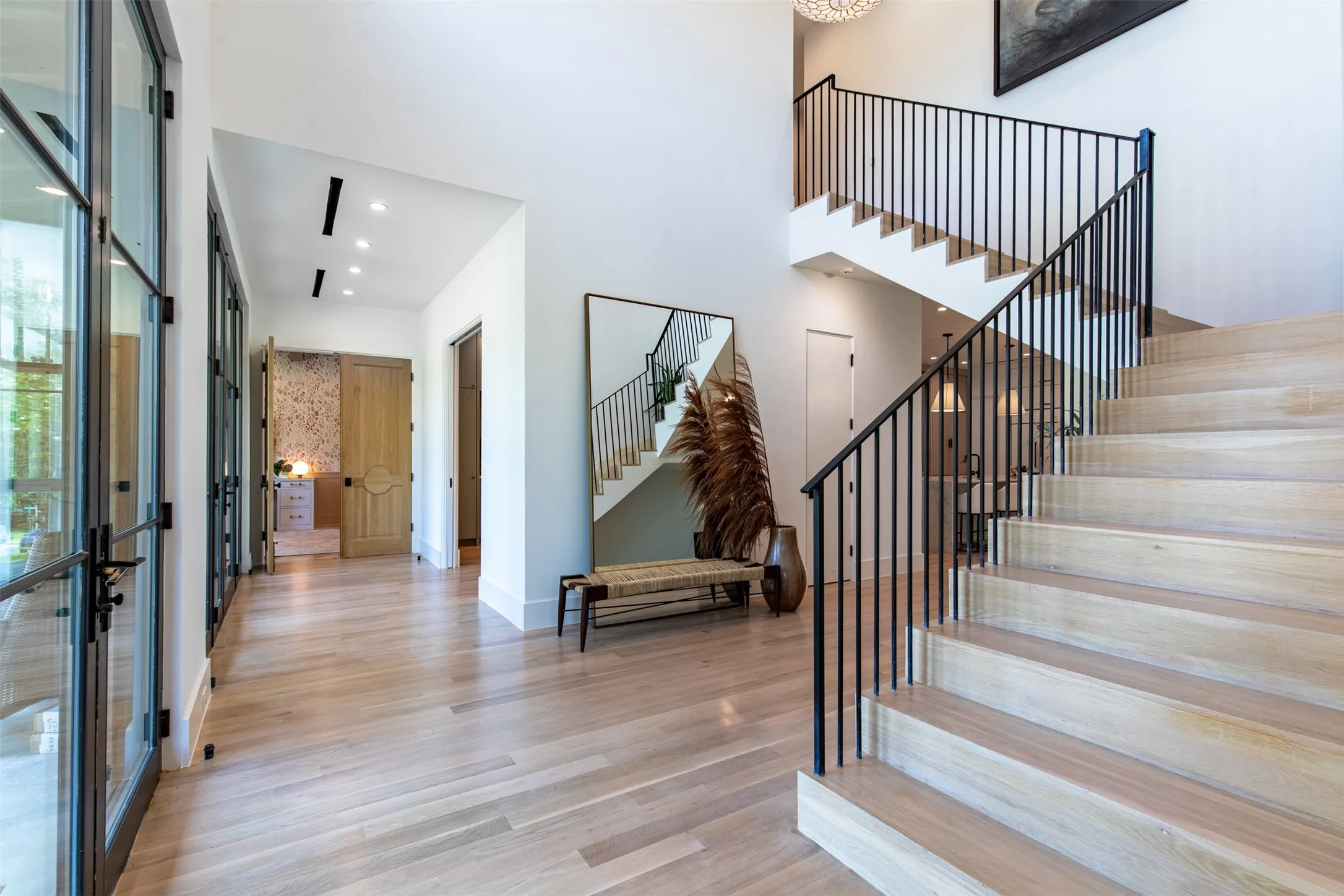 Light wood-finish flooring extending through a spacious entryway featuring an open staircase with black iron railings, a wall of glass-paned doors, recessed lighting, and a chandelier