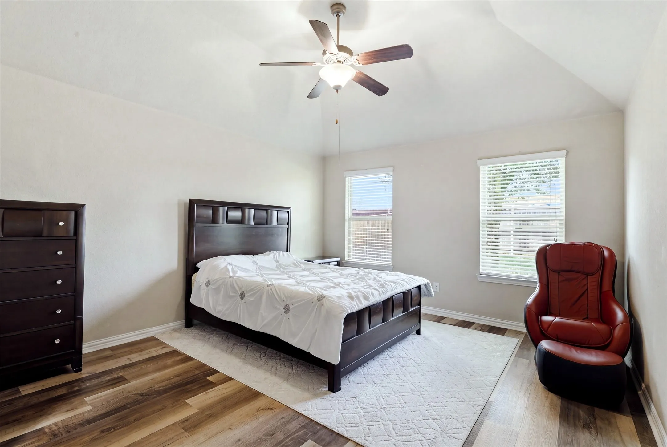 Vaulted ceiling, ceiling fan with integrated lighting, wood-finish flooring, and multiple windows with blinds