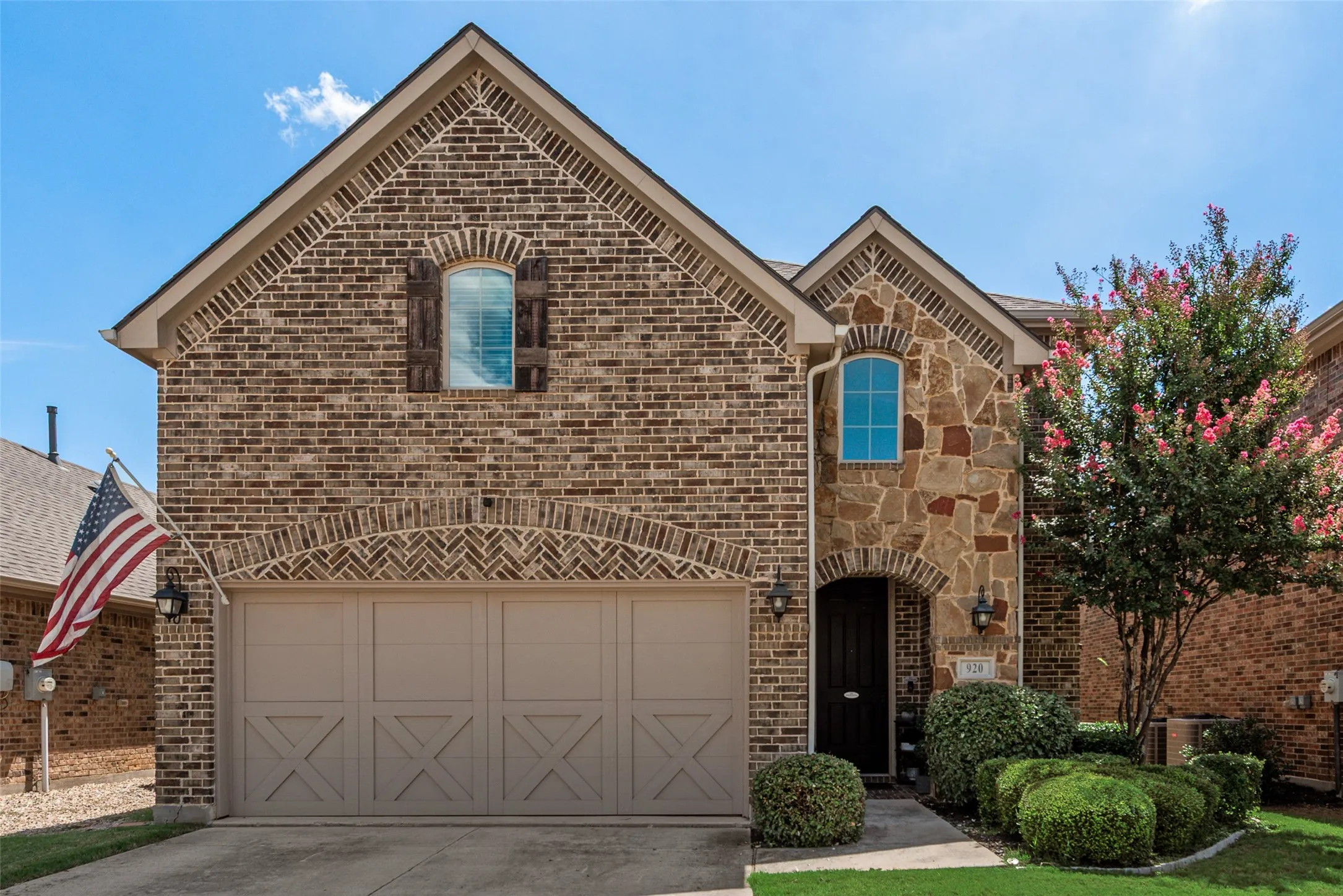 Brick and stone facade featuring an arched entryway, decorative garage door, and mature landscaping