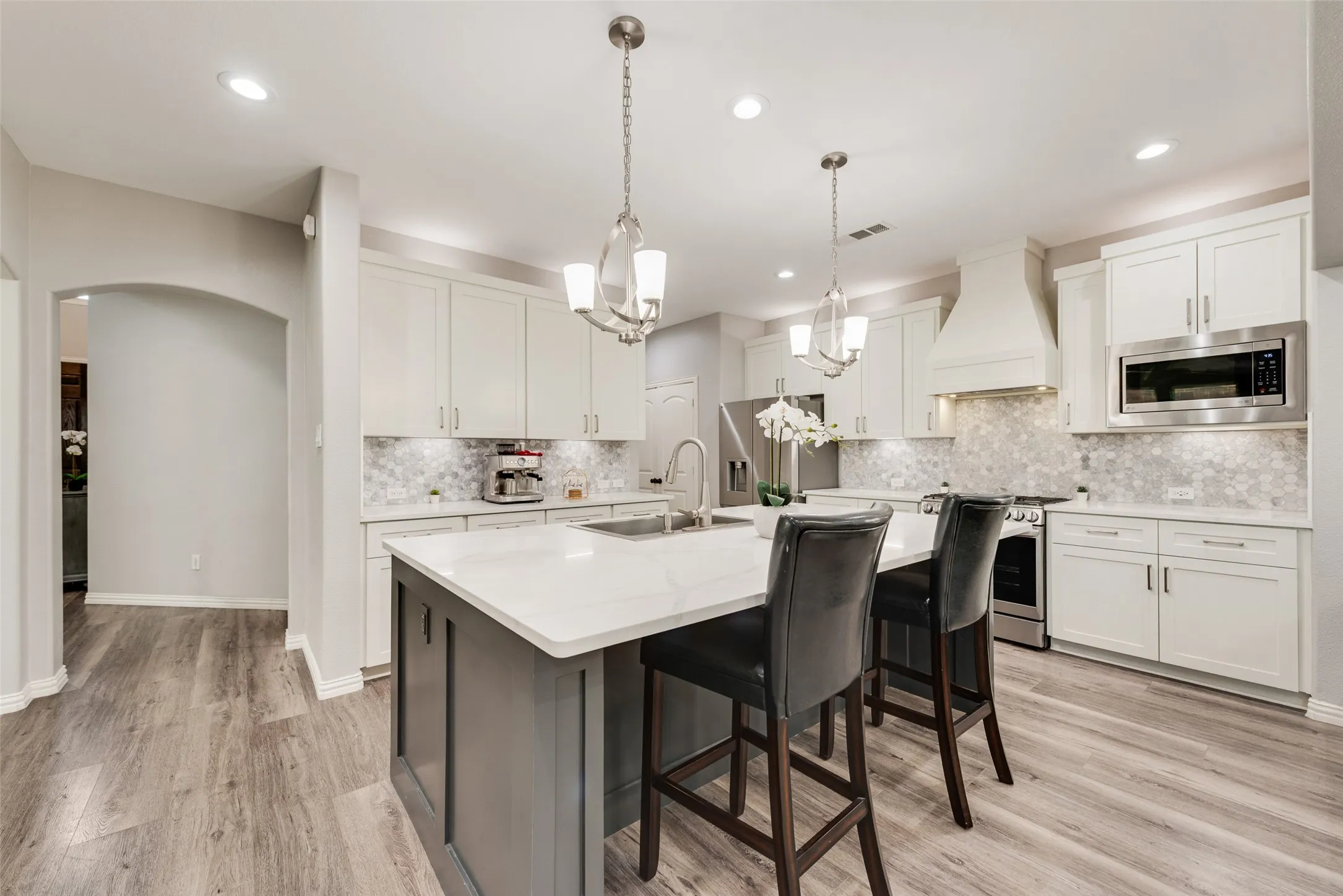 Spacious kitchen featuring an expansive island with seating, white shaker cabinetry, stainless steel appliances, a white mosaic tile backsplash, and wood-finish flooring