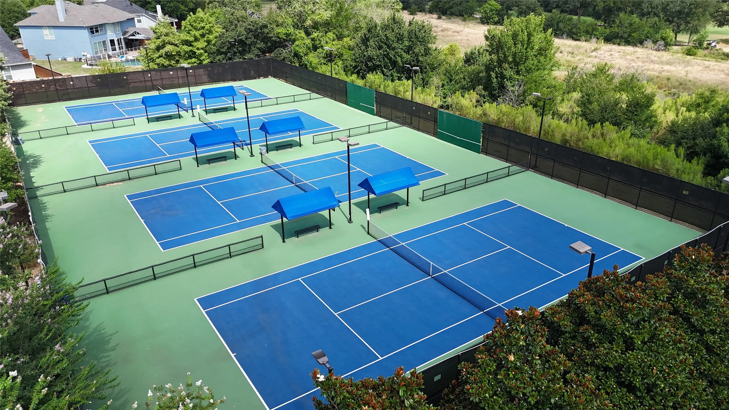 Multiple blue tennis courts with green surrounds, black fencing, and covered seating areas
