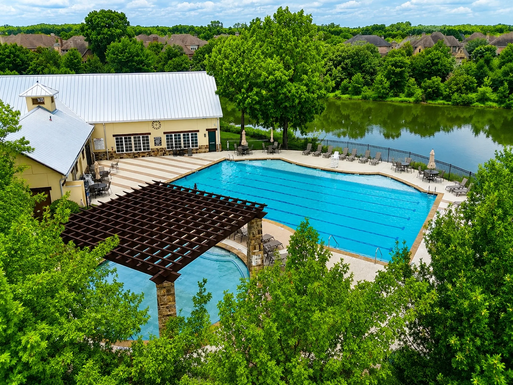 Outdoor swimming pool complex featuring a large rectangular pool with lane markers, an adjacent smaller pool under a dark wood pergola, a cream-colored building with a light-colored metal roof, and a paved patio area with outdoor seating, all bordering a body of water