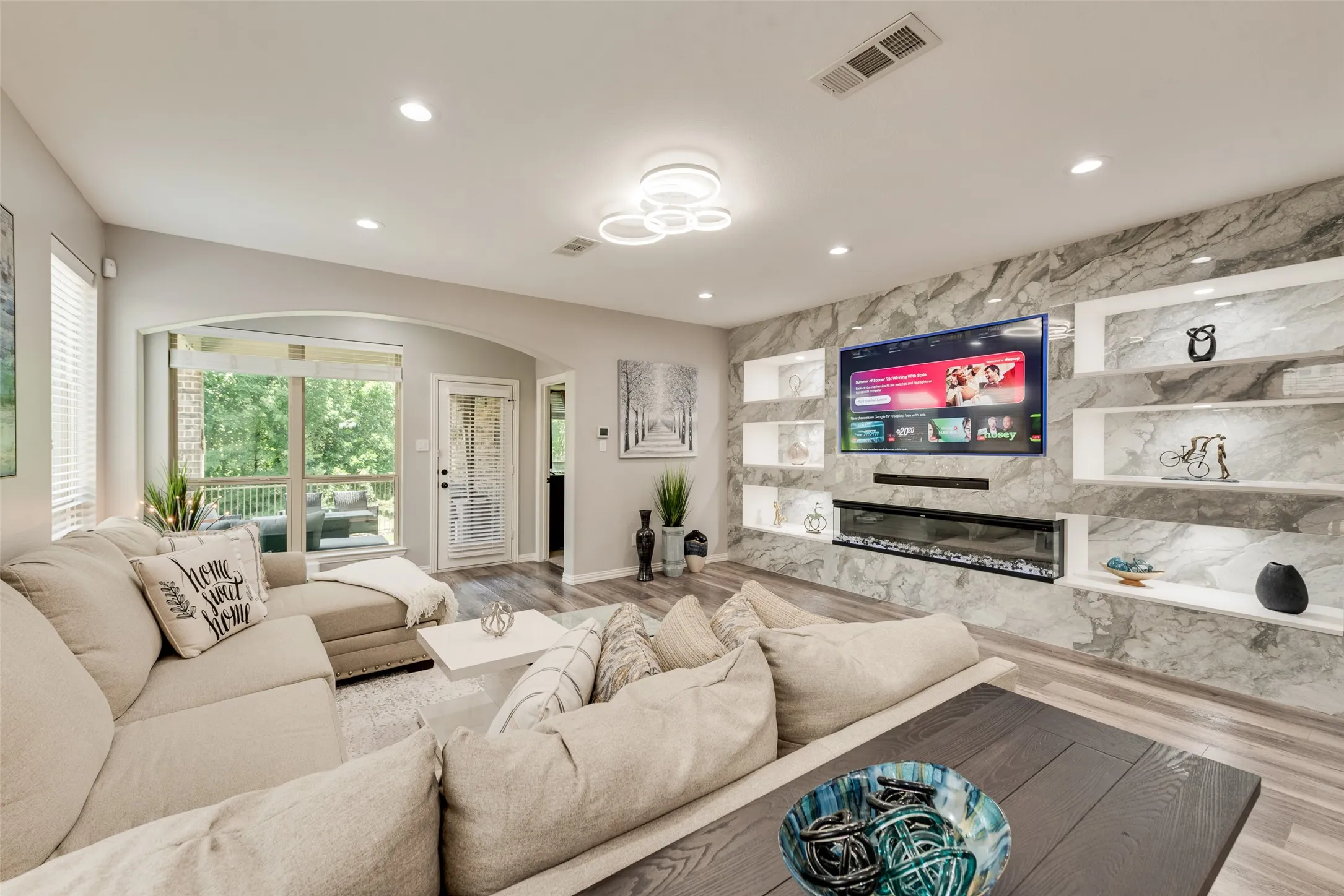 Living area featuring a stone-look accent wall, built-in shelving with integrated lighting, a linear electric fireplace, wood-finish flooring, and recessed ceiling lights
