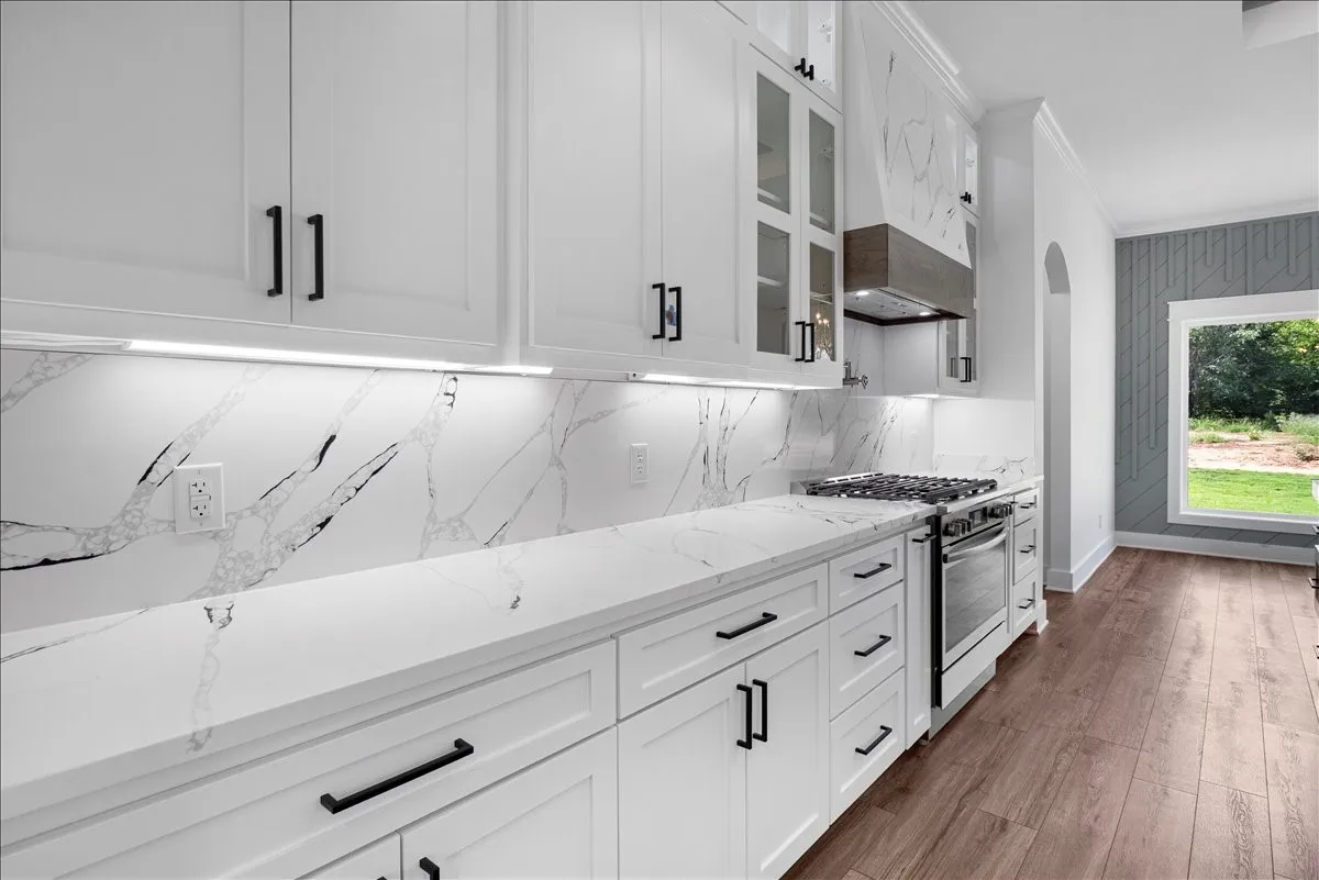 White cabinetry with matte black hardware, integrated range with a stainless steel oven, white countertops and backsplash, under-cabinet lighting, and wood-finish flooring