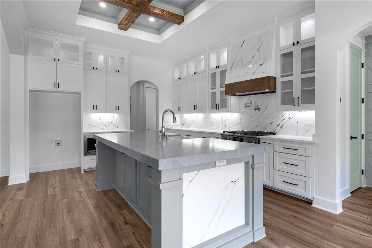 Gourmet kitchen featuring a large central island with a waterfall edge, extensive white cabinetry with glass-front uppers, a gas range with a wood-accented vent hood, and wood-finish flooring