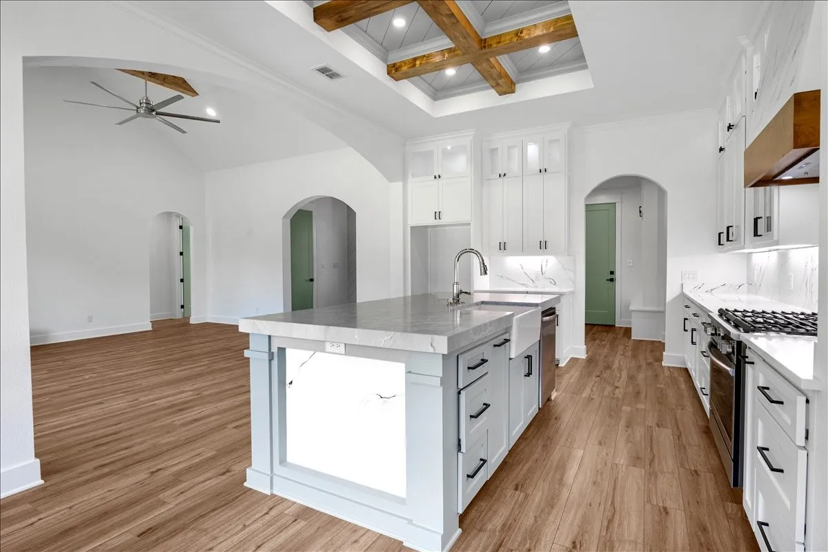 Kitchen featuring a central island with an integrated sink, white cabinetry with dark hardware, a gas range, wood-finish flooring, and a tray ceiling with exposed wood beams