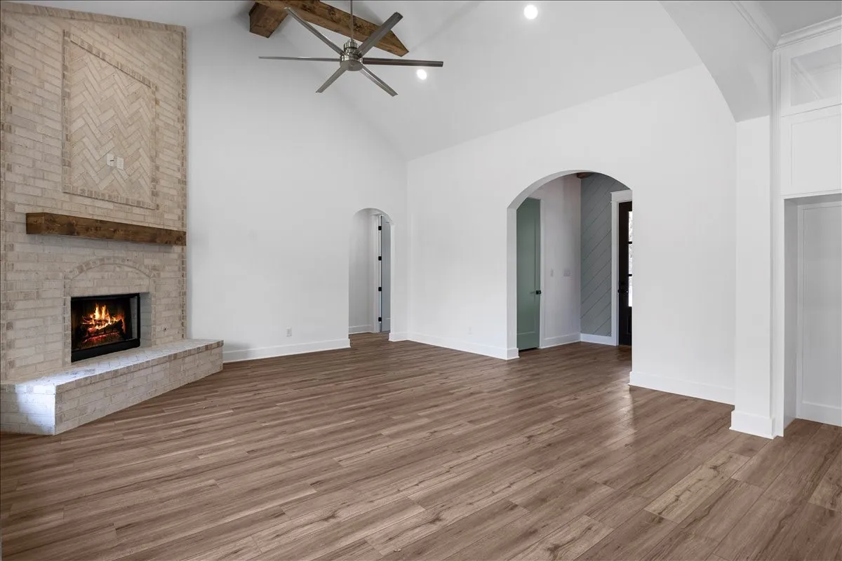 Vaulted ceiling with exposed wood beams, wood-finish flooring, brick fireplace with a wood mantel, arched doorway, and recessed lighting