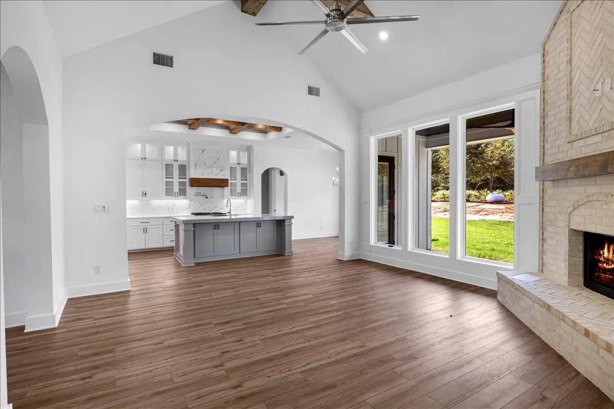 Vaulted ceilings with exposed wood beams, wood-finish flooring, a brick fireplace, a kitchen island with a built-in sink, and large windows