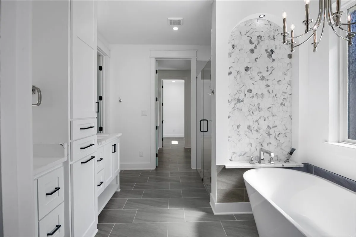 Freestanding soaking tub, walk-in shower with glass enclosure, arched tiled accent wall, white cabinetry with matte black hardware, and large format floor tiling