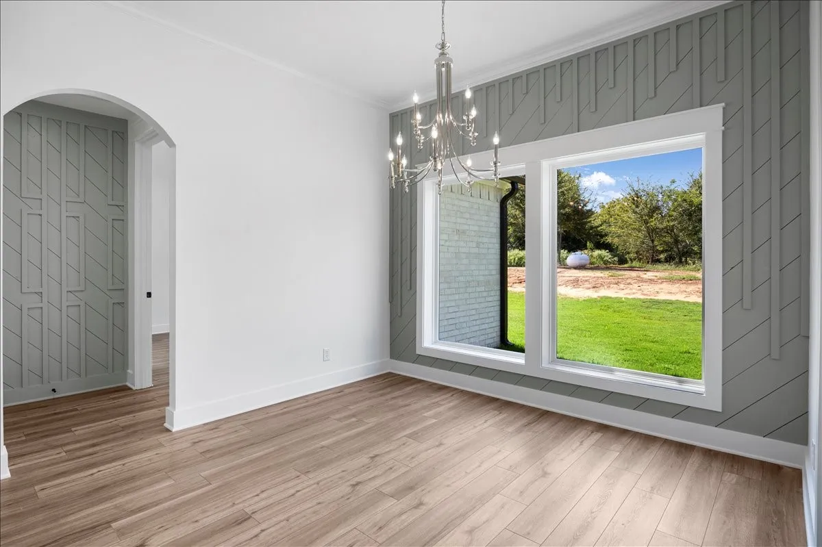 Wood-finish flooring, decorative wall paneling, arched doorway, chandelier, and a pair of large windows