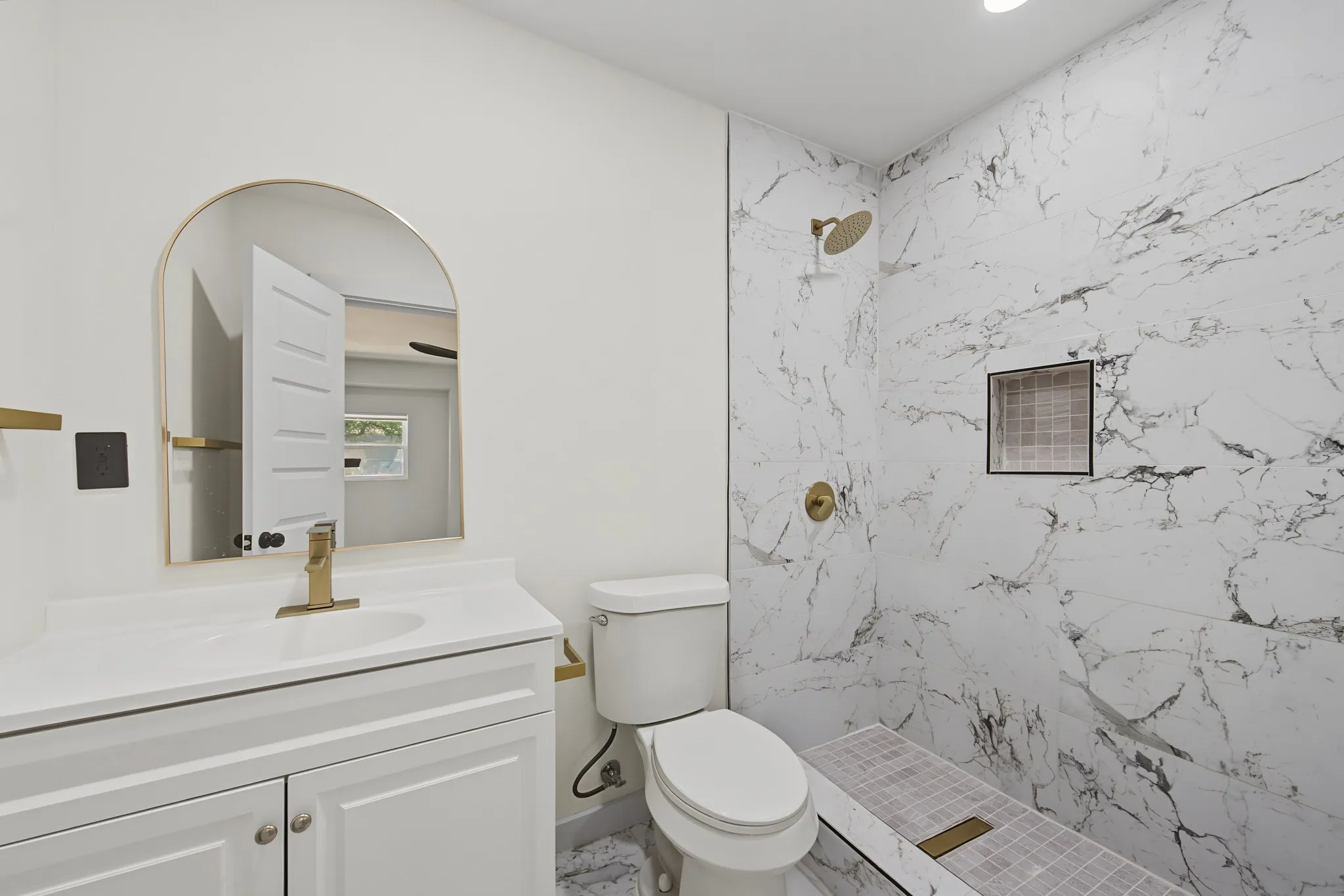 Bathroom featuring a white vanity with a rectangular sink, a gold-framed arched mirror, a toilet, a shower with large format tile walls, and a built-in shower niche