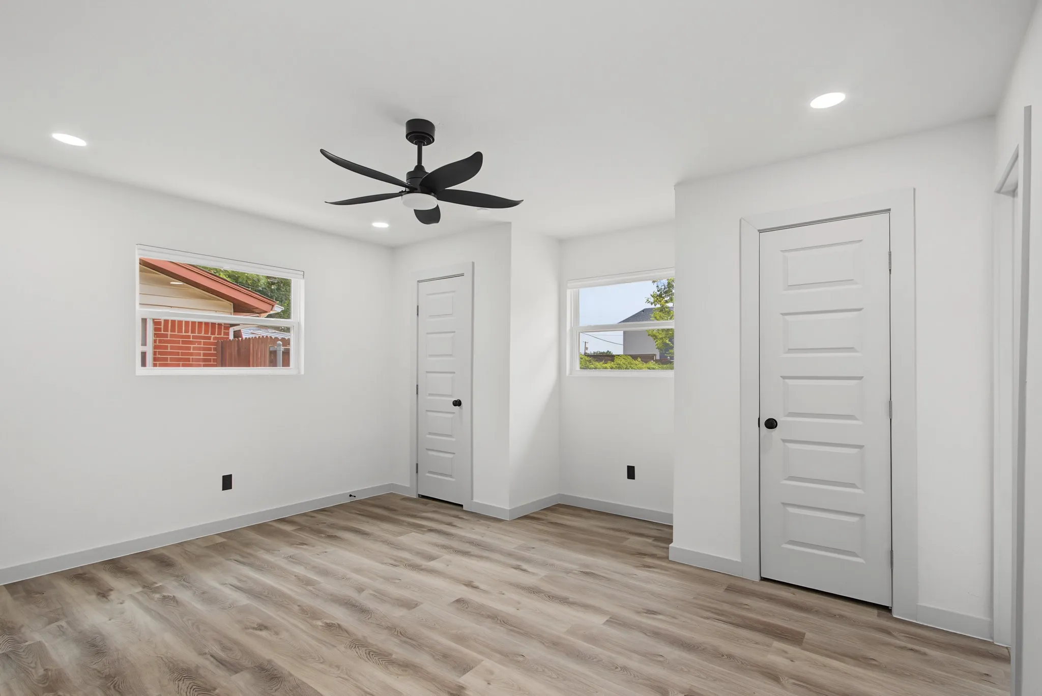 Spacious interior featuring wood-finish flooring, bright white walls, two windows, two paneled doors, and a ceiling fan with integrated lighting