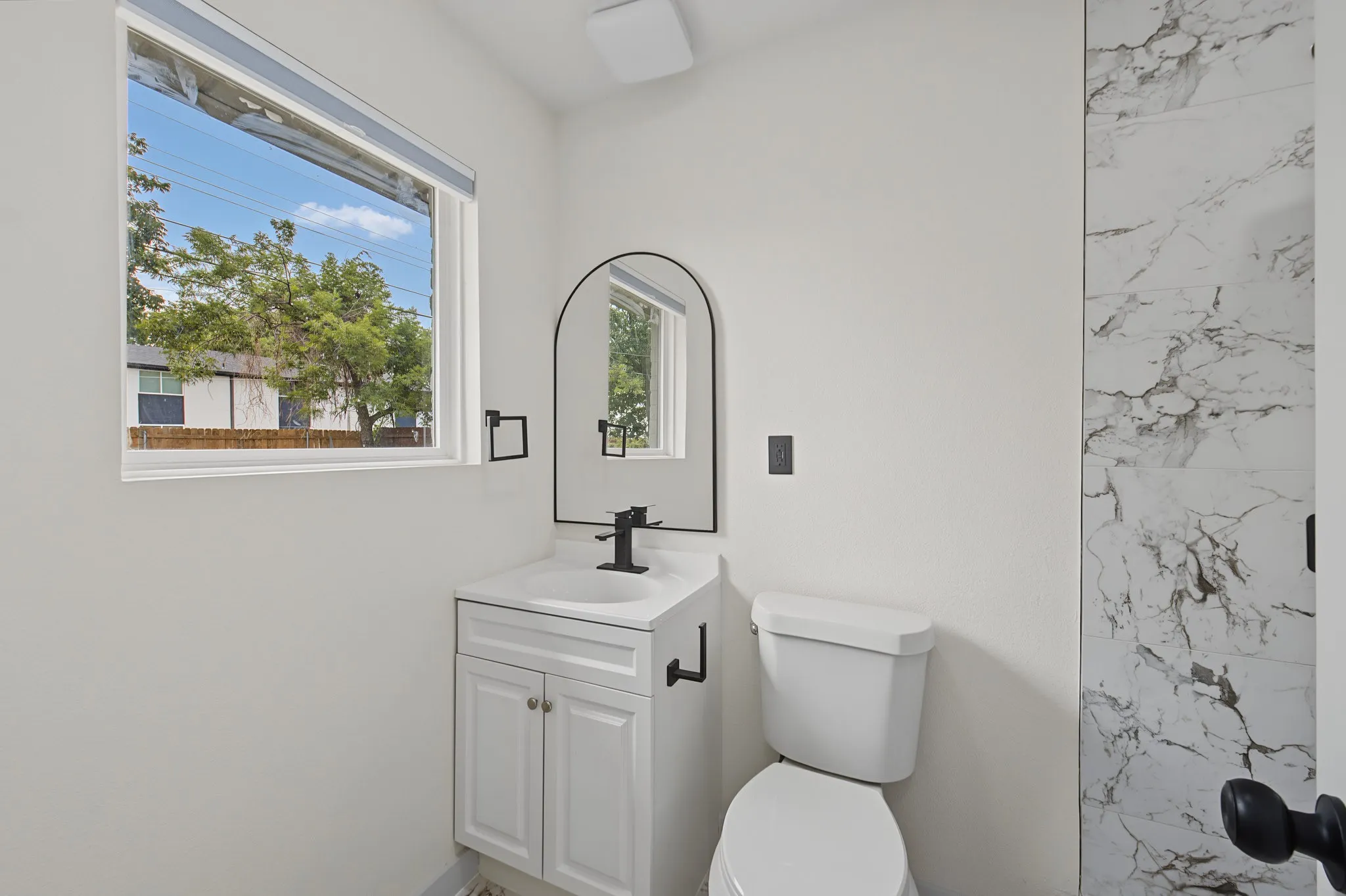 Bathroom featuring a single vanity with a white countertop and cabinet storage, a matte black arched mirror, a toilet, and a shower with large format marble-look tile