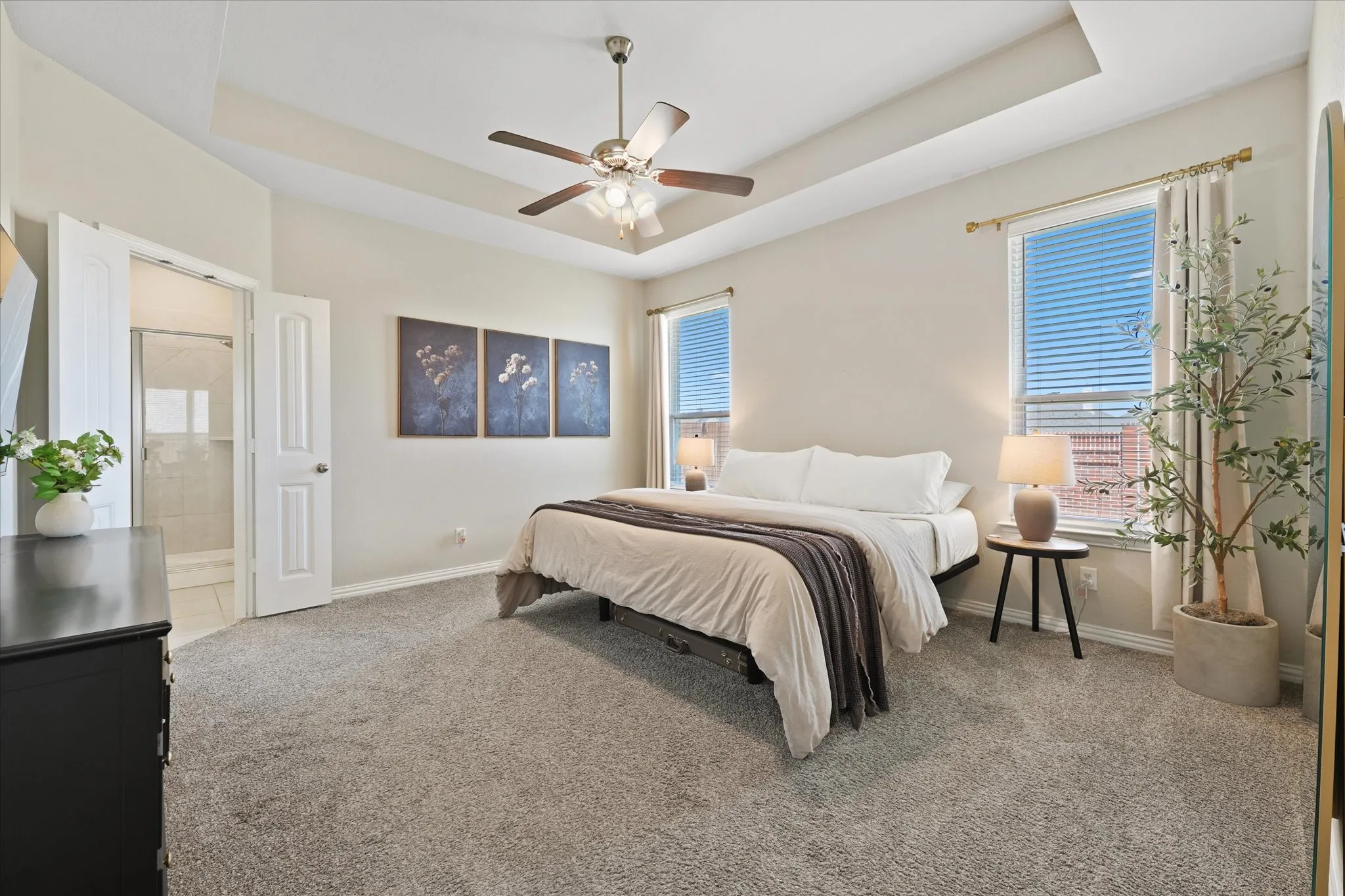 Bedroom featuring tray ceiling, ceiling fan, carpet flooring, dual windows with blinds, and an adjoining bathroom with a glass-enclosed shower