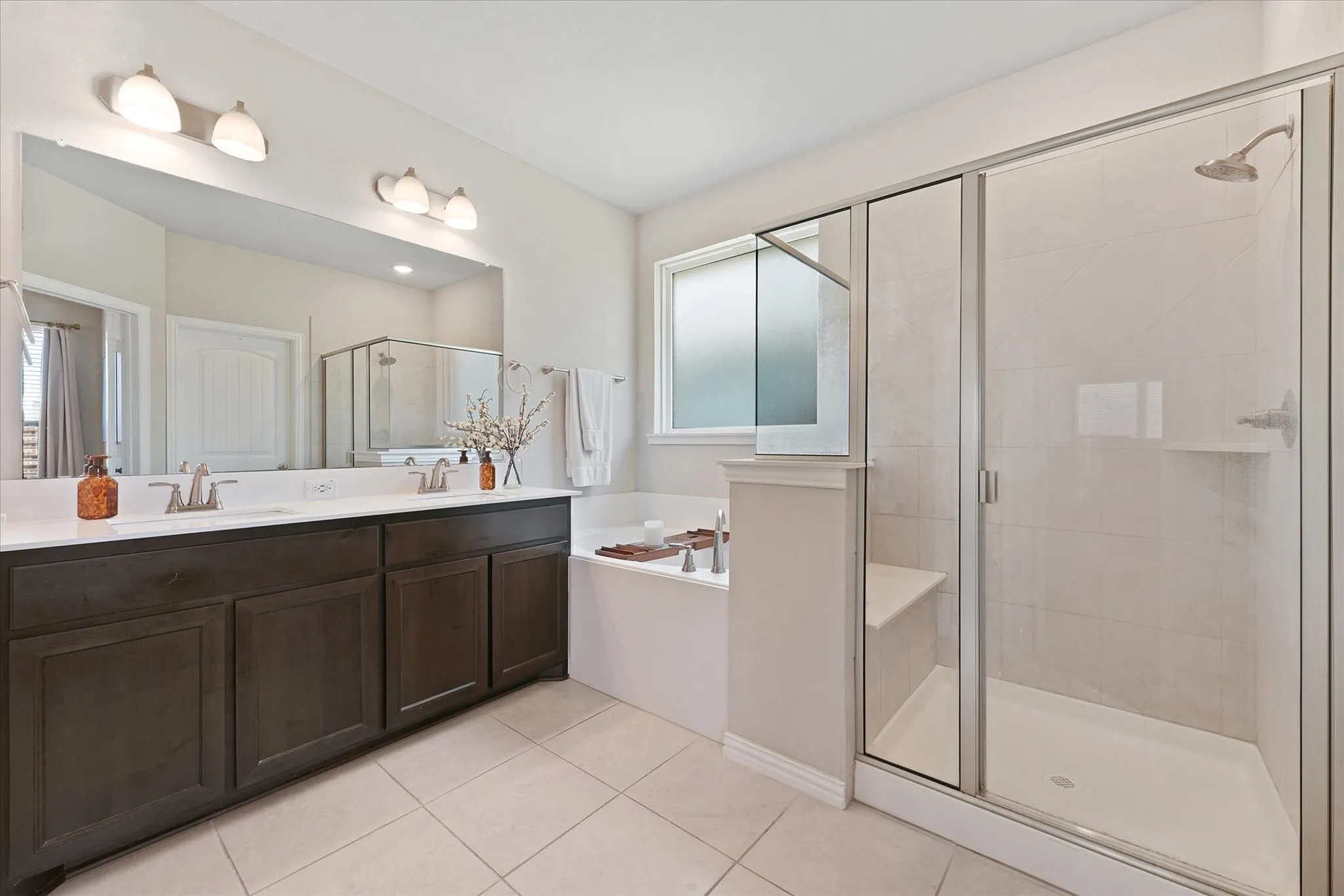 Spa-inspired primary bathroom featuring a dark wood vanity with dual sinks, extensive mirror with integrated lighting, and a spacious walk-in shower with frameless glass enclosure and tile surround